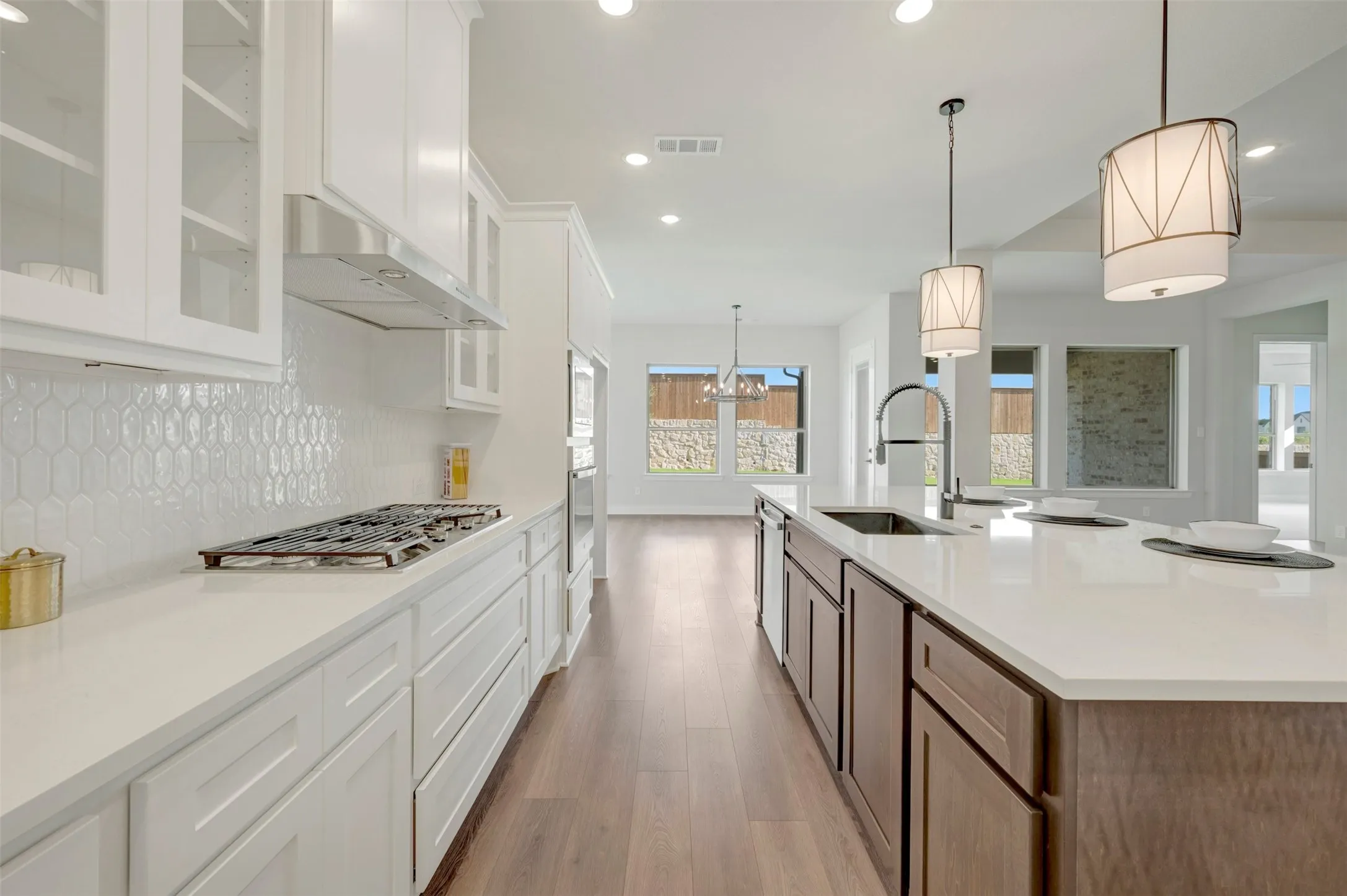 Modern kitchen featuring white shaker cabinetry, an elongated hexagonal tile backsplash, a gas cooktop with stainless steel range hood, and a large central island with a sink and pendant lighting