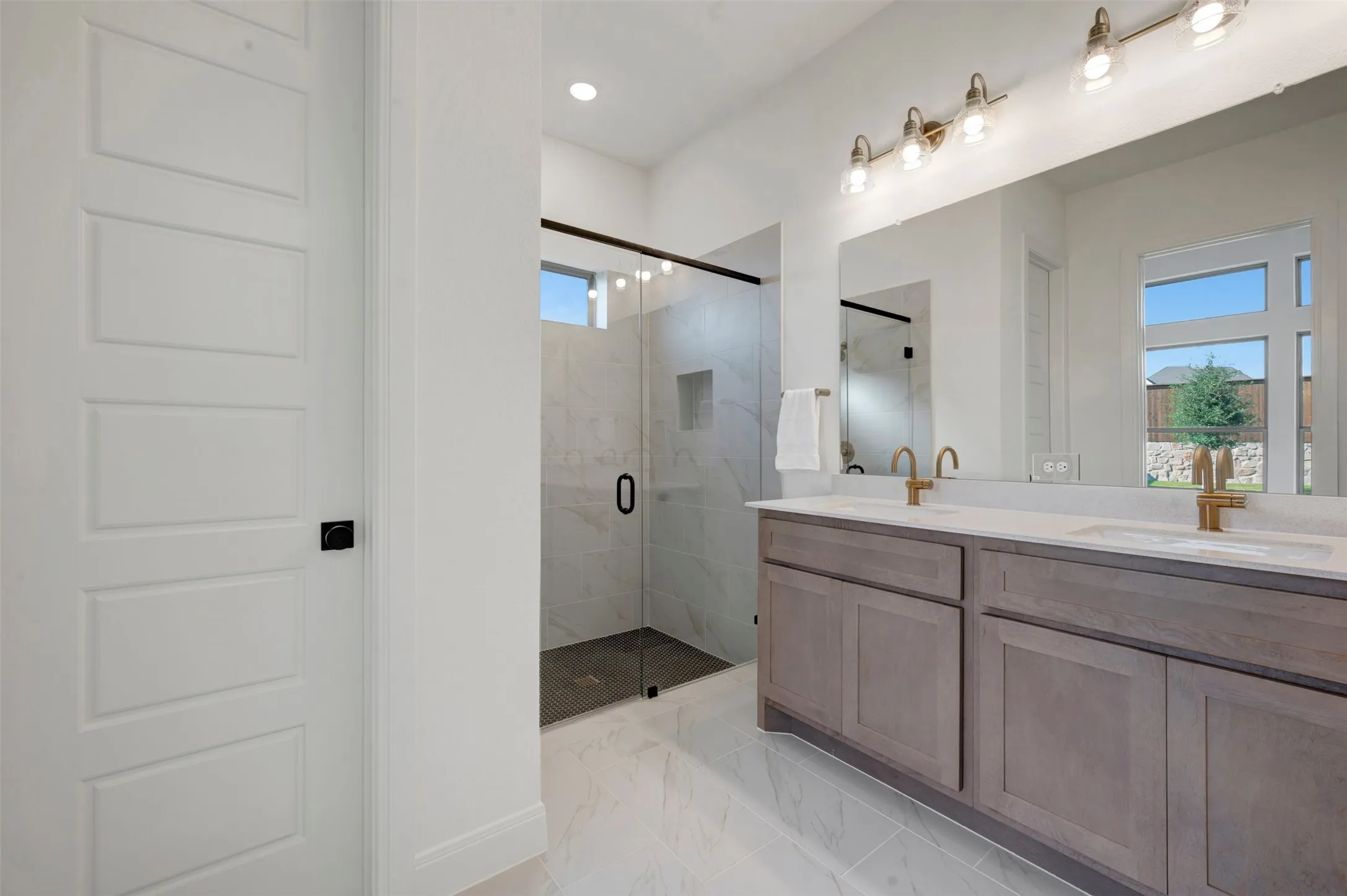 Spacious bathroom featuring a dual vanity with light wood-finish cabinetry and light countertops, a large mirror, a frameless glass shower enclosure with floor-to-ceiling tile, and light tile flooring