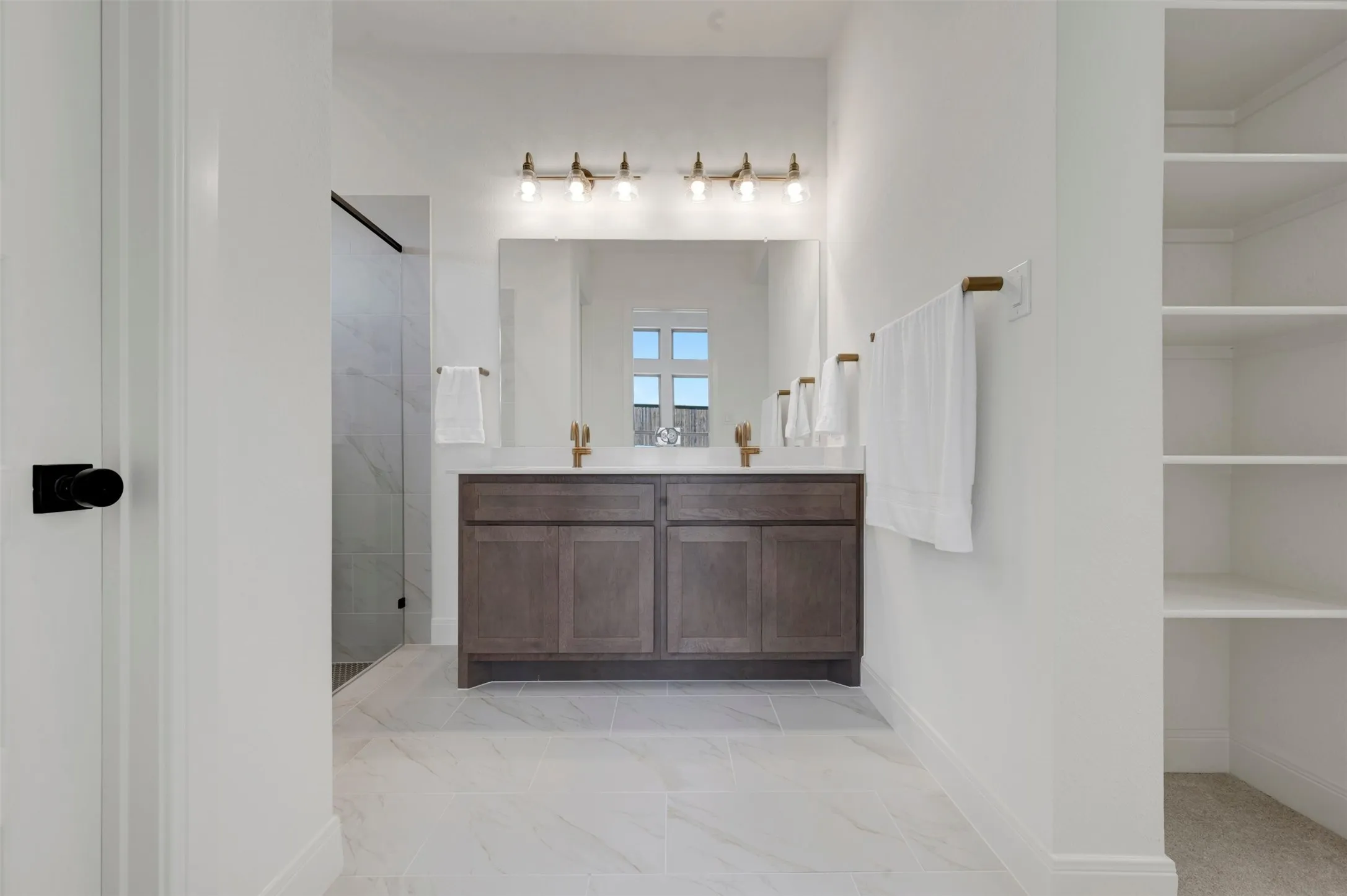 Bathroom featuring a dual vanity with a wood-finish cabinet, brushed gold fixtures, a glass-enclosed shower with large format tile, and built-in shelving