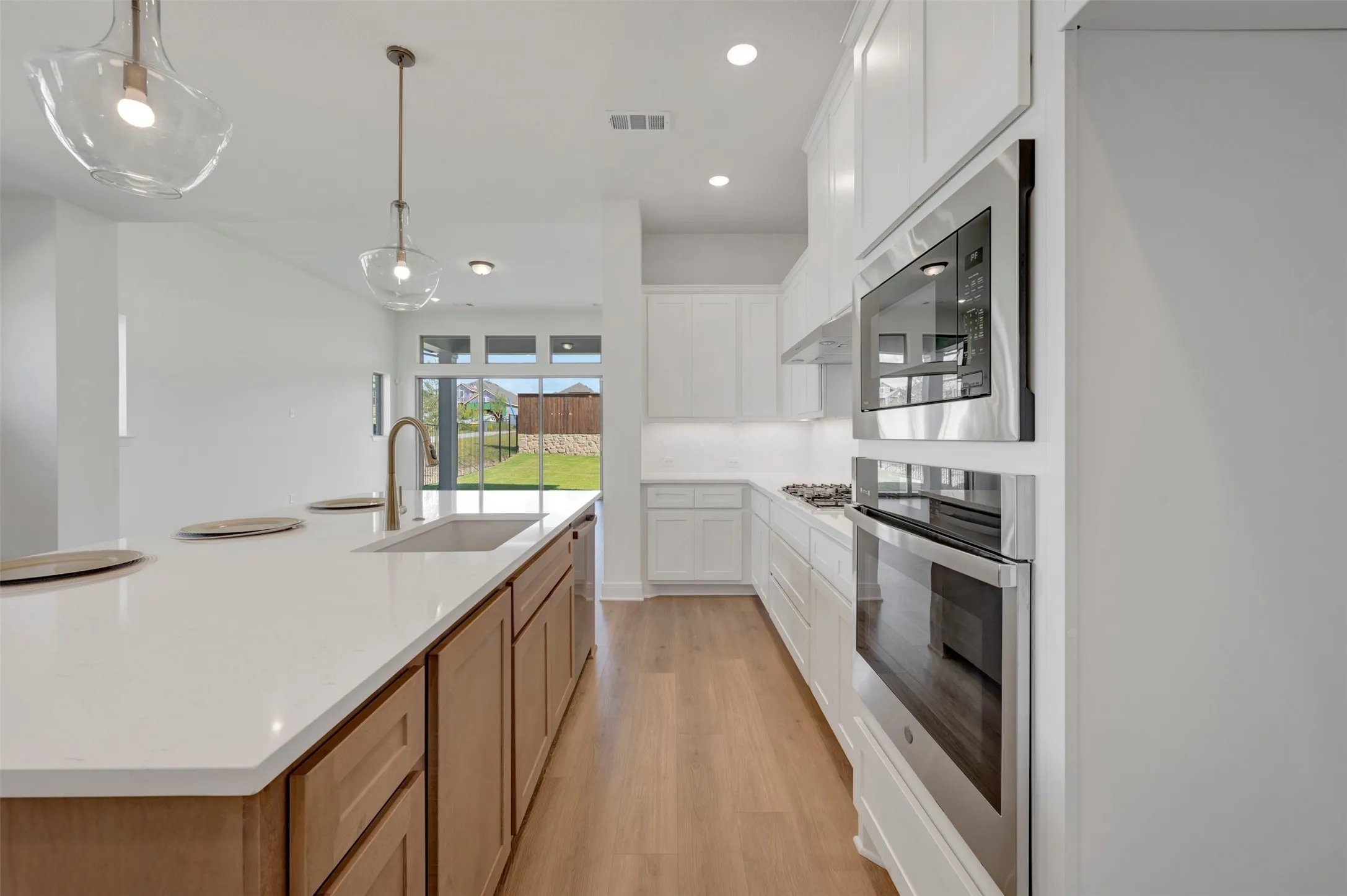 Open-concept kitchen featuring an island with a built-in sink and gold-finish faucet, light wood-finish flooring, white upper and lower cabinetry, stainless steel built-in microwave and oven, and recessed lighting
