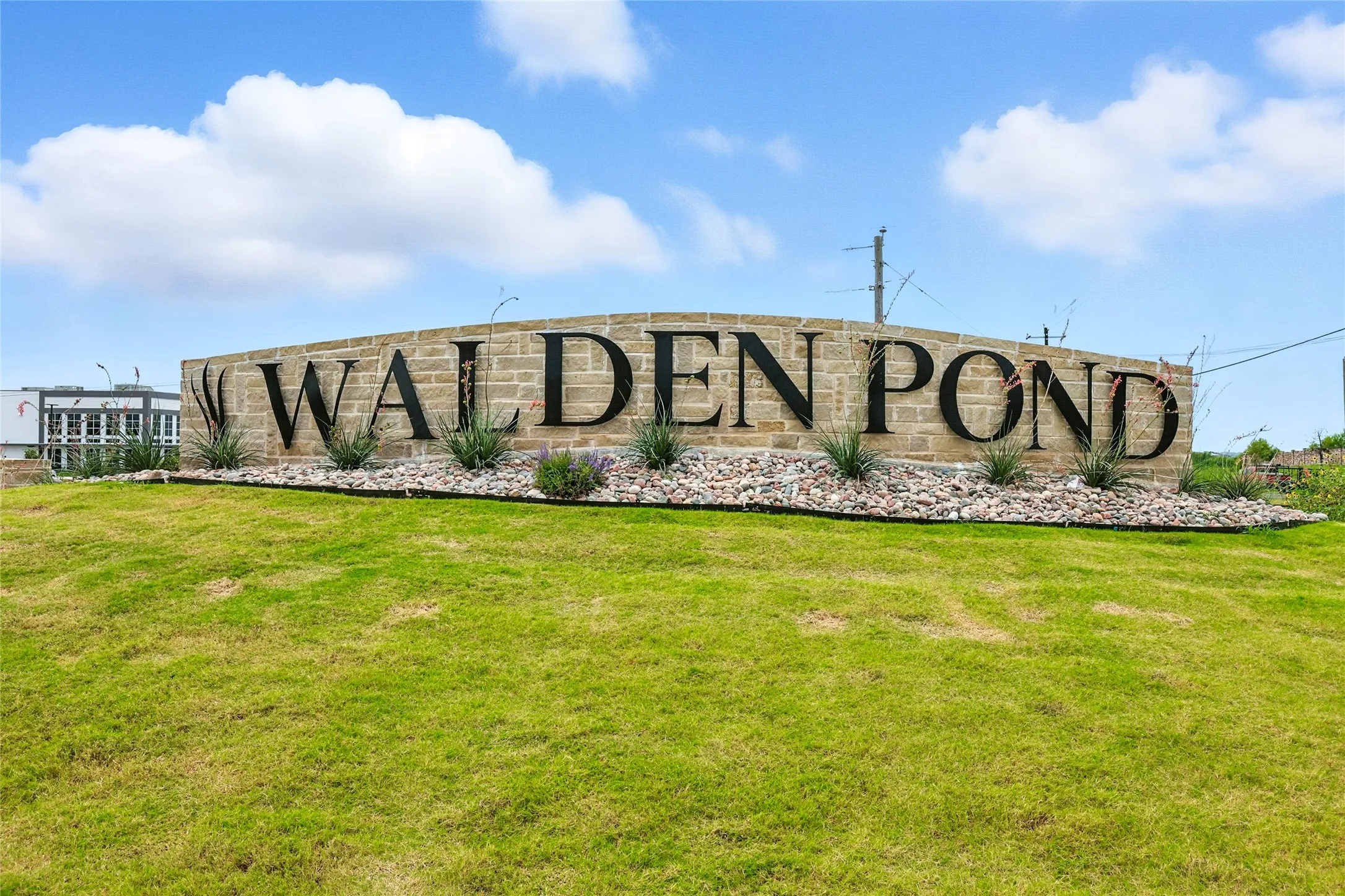 Stone entrance monument with integrated dark metal lettering for "WALDEN POND," set against a manicured green lawn with decorative rock landscaping and planted shrubs