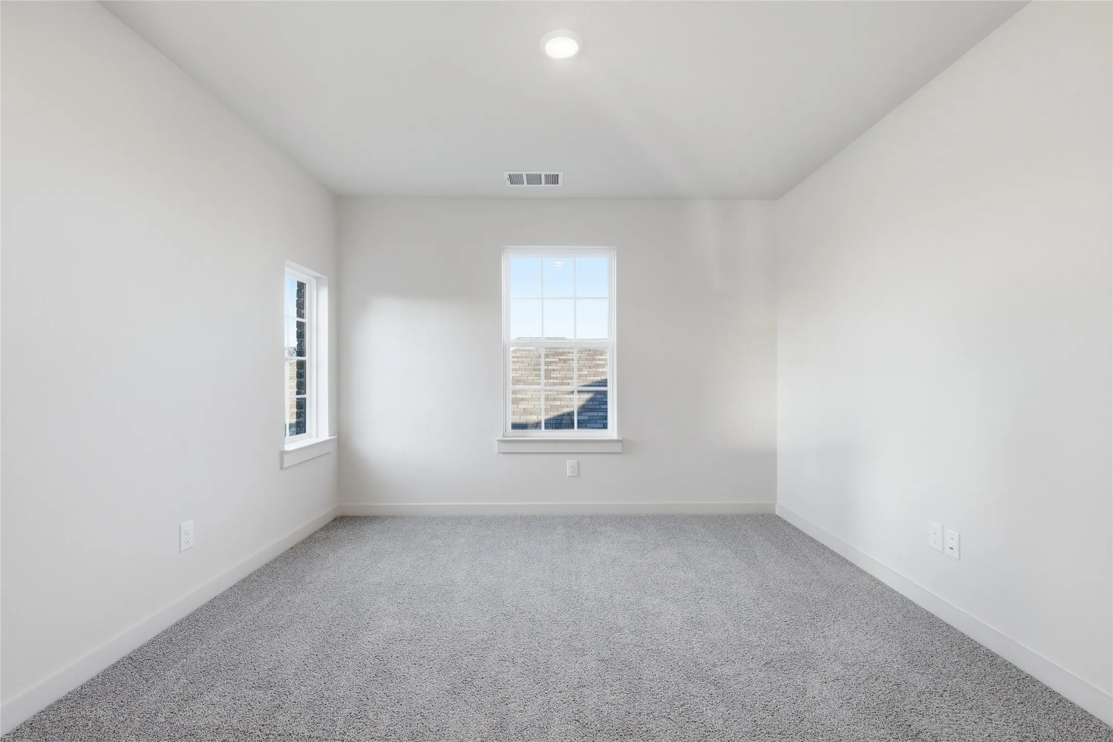 Spacious room featuring light gray carpeting, neutral wall paint, two windows with white trim, and a recessed ceiling light