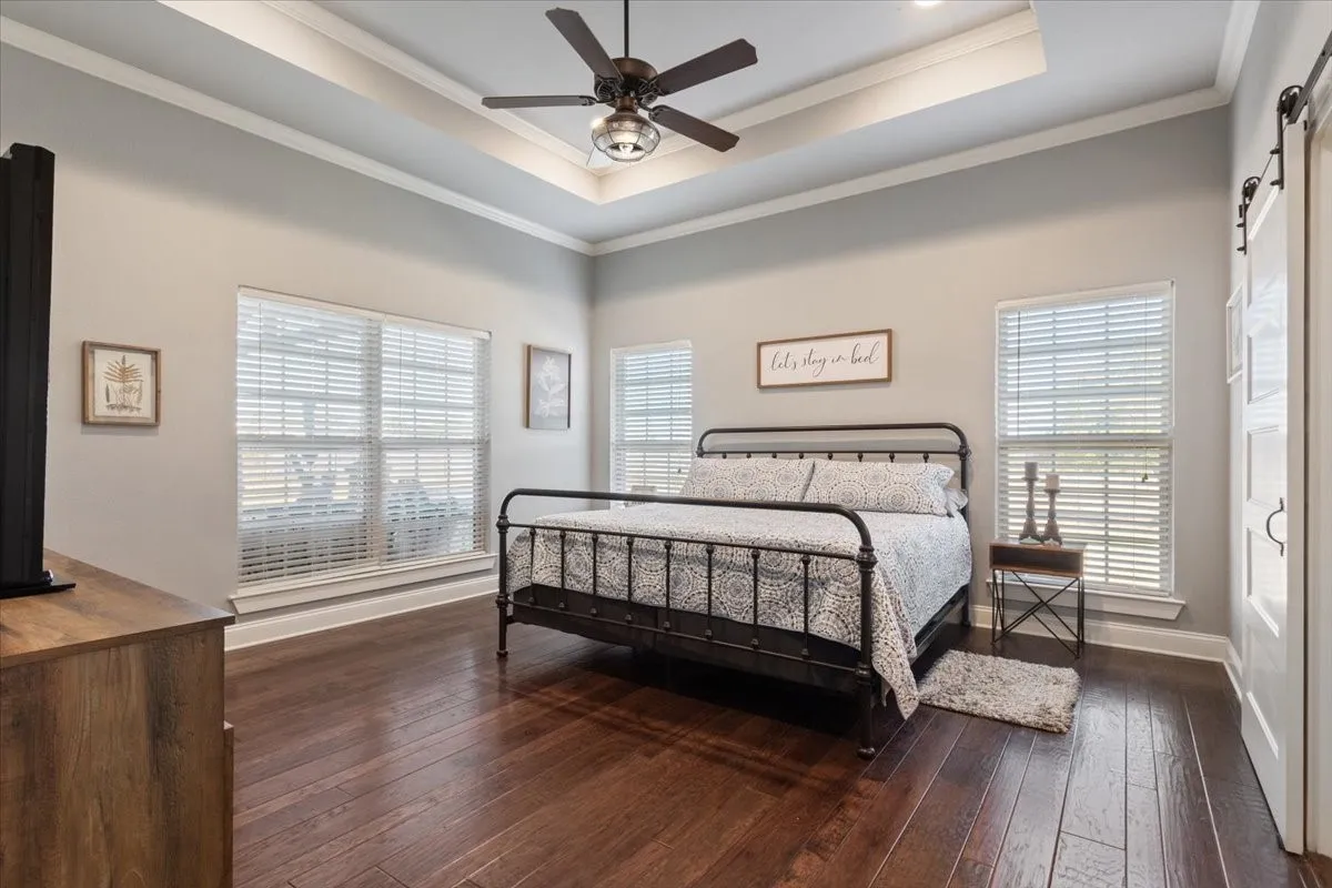Bedroom featuring a barn door, crown molding, dark wood-style floors, ceiling fan, and a tray ceiling