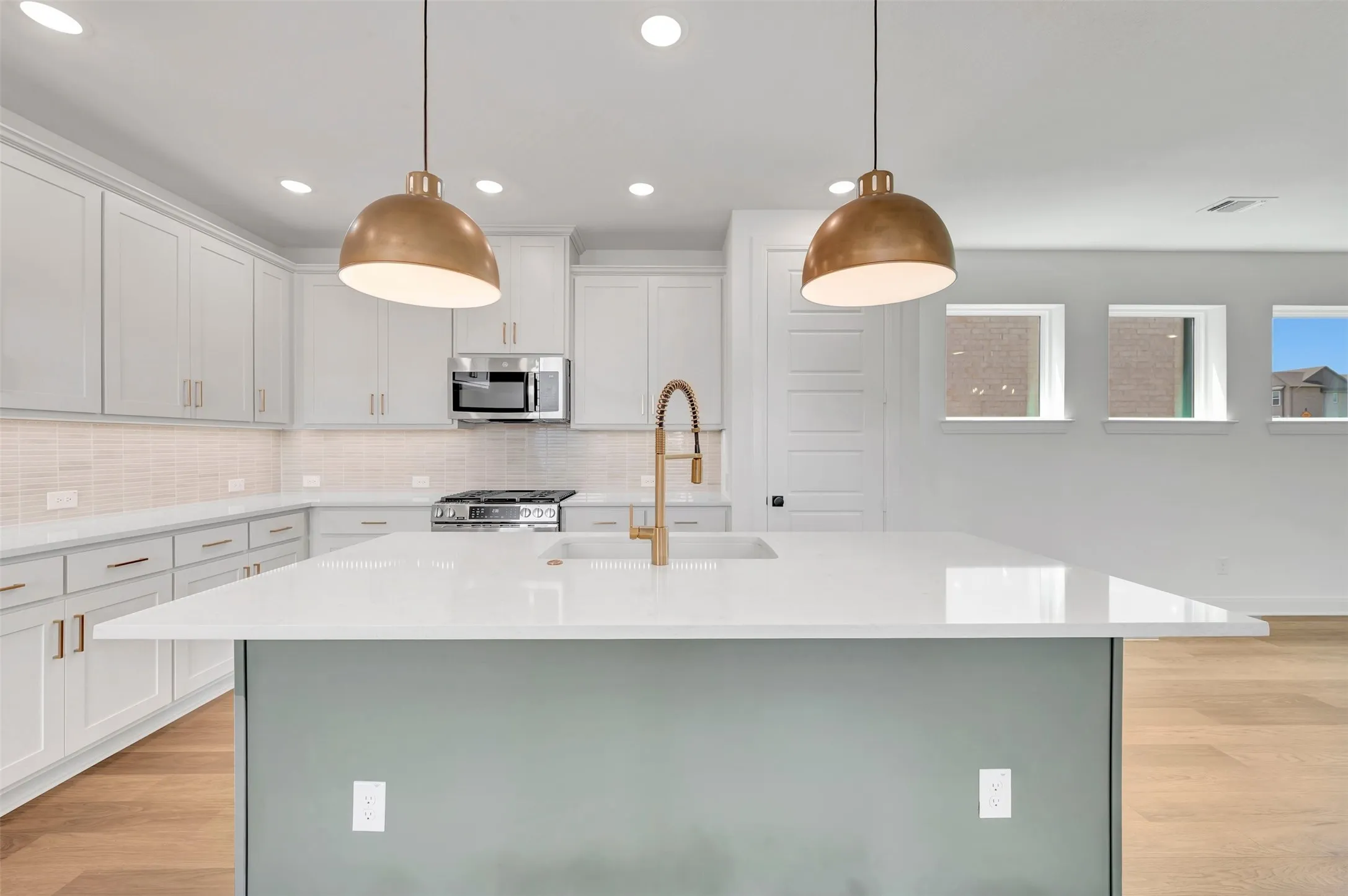 Spacious kitchen featuring a large island with a light-colored countertop, white cabinetry with gold-toned hardware, a stainless steel gas range, and two suspended gold-toned pendant lights