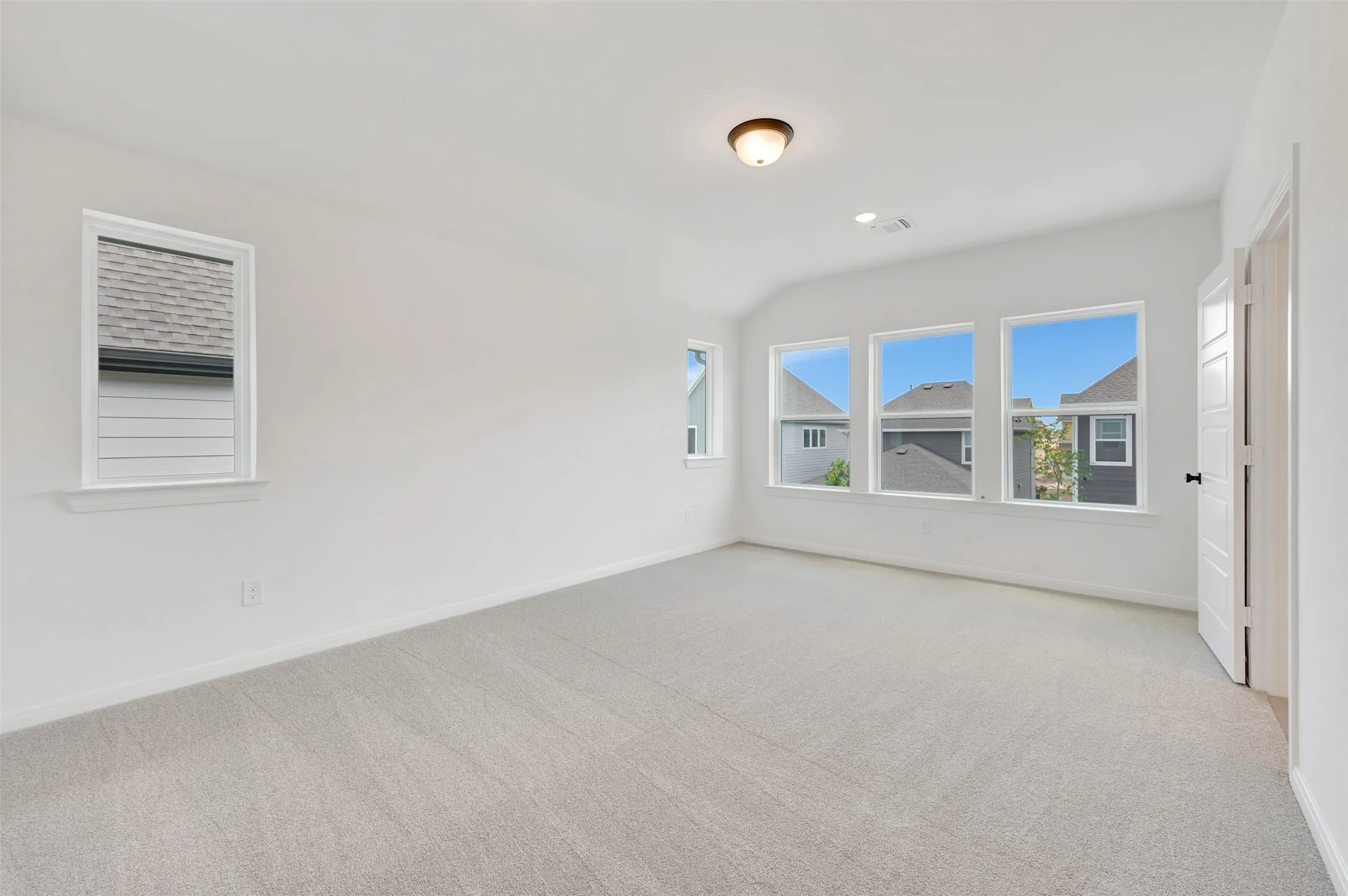 Spacious room featuring light-colored carpeting, multiple white-framed windows, a white paneled door with black hardware, a flush-mount ceiling light fixture, and recessed lighting
