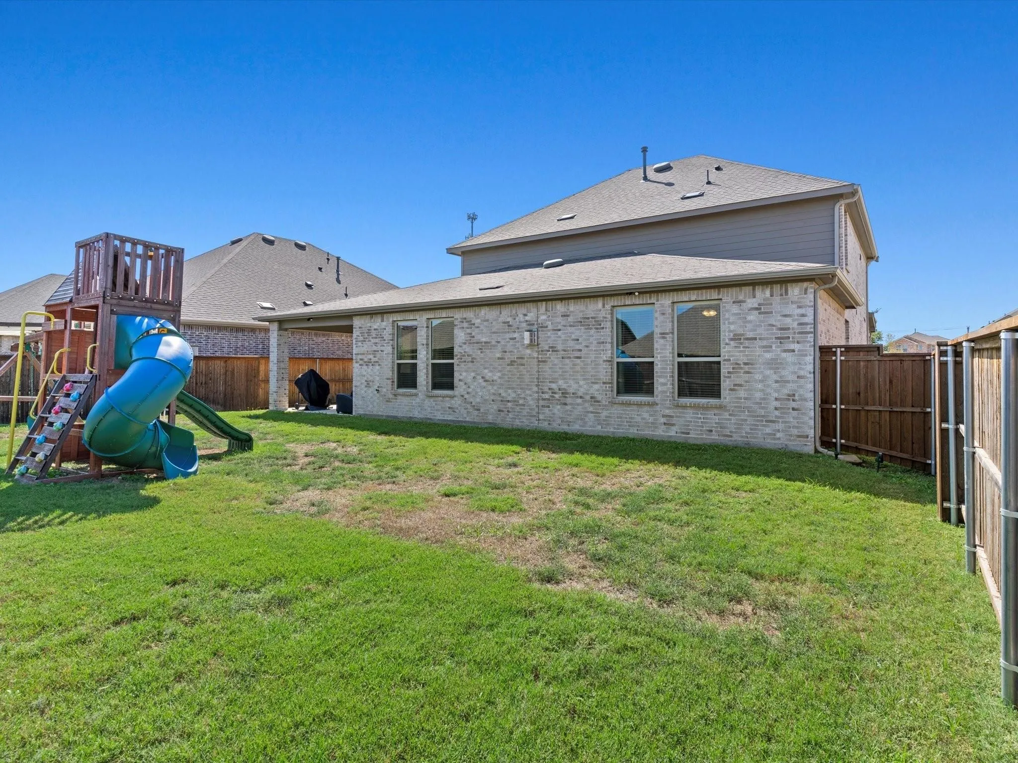 Expansive backyard featuring a manicured lawn, brick exterior, covered patio, multiple windows, and a privacy fence