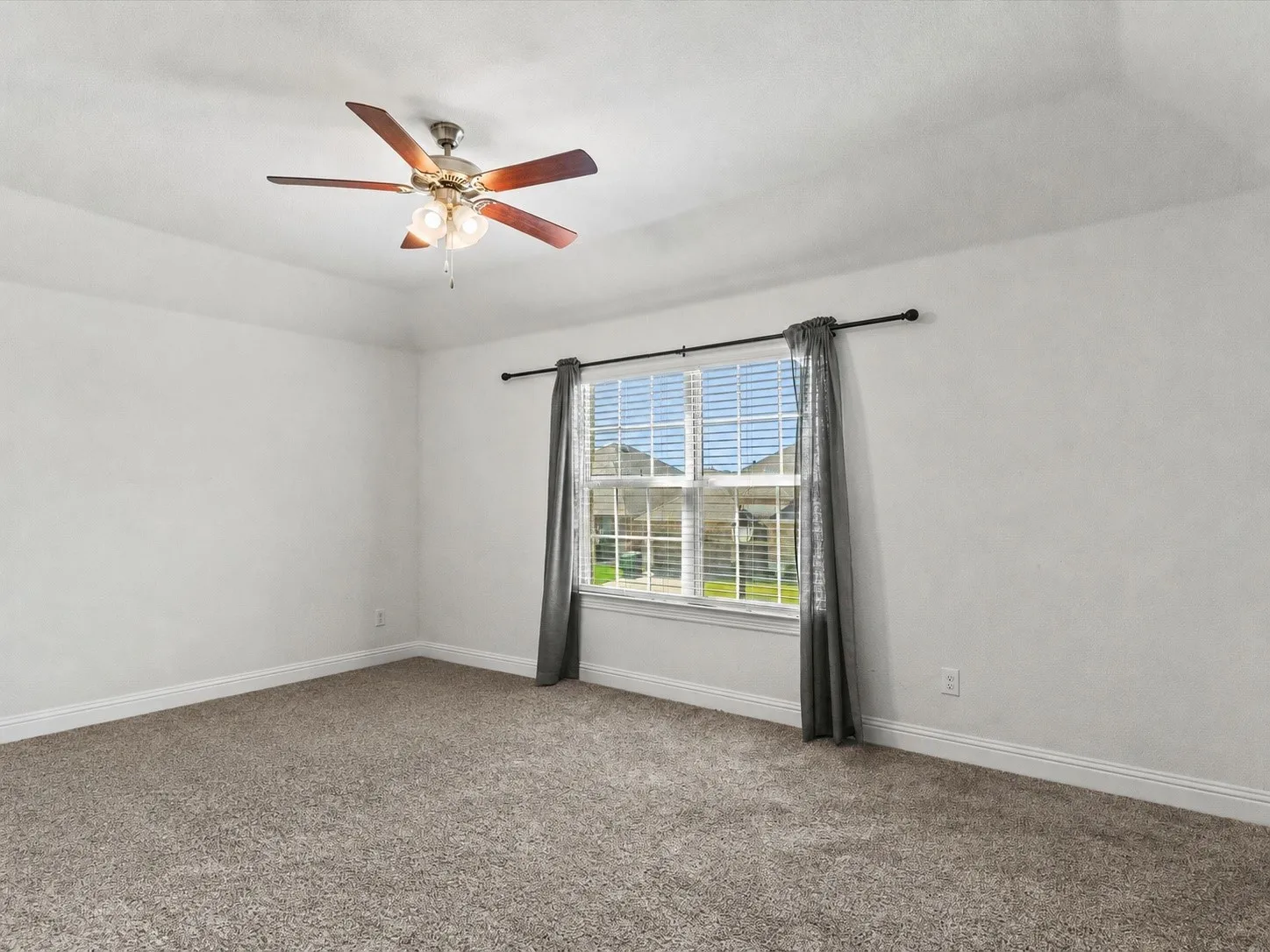 Spacious room featuring neutral carpeting, white walls, a large window with horizontal blinds, a ceiling fan with integrated lighting, and a white trim baseboard