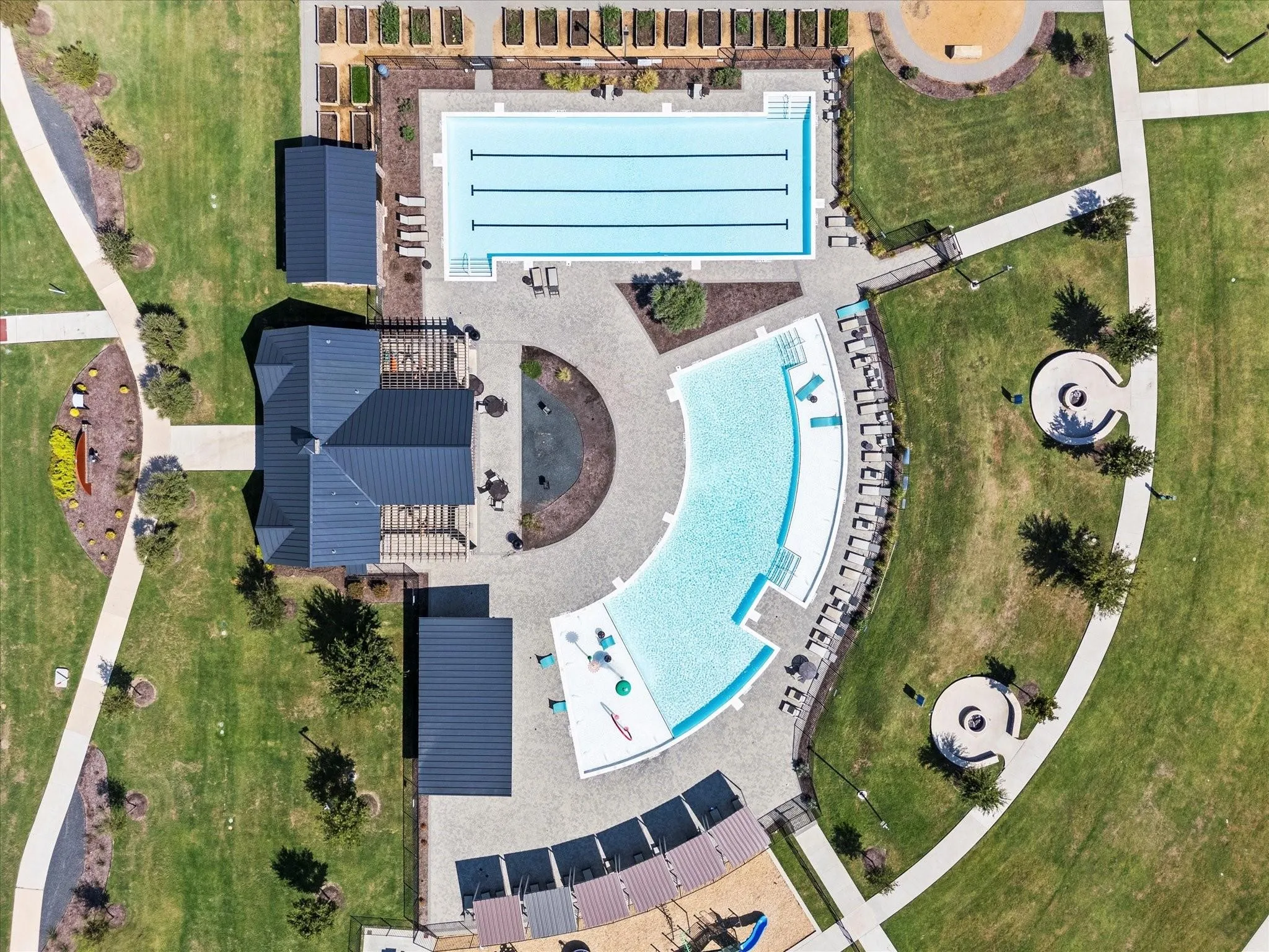 Aerial perspective of a community amenity center featuring a rectangular lap pool, a curved leisure pool, a central clubhouse structure with dark roofing, multiple shaded seating areas, and surrounding green spaces with walking paths