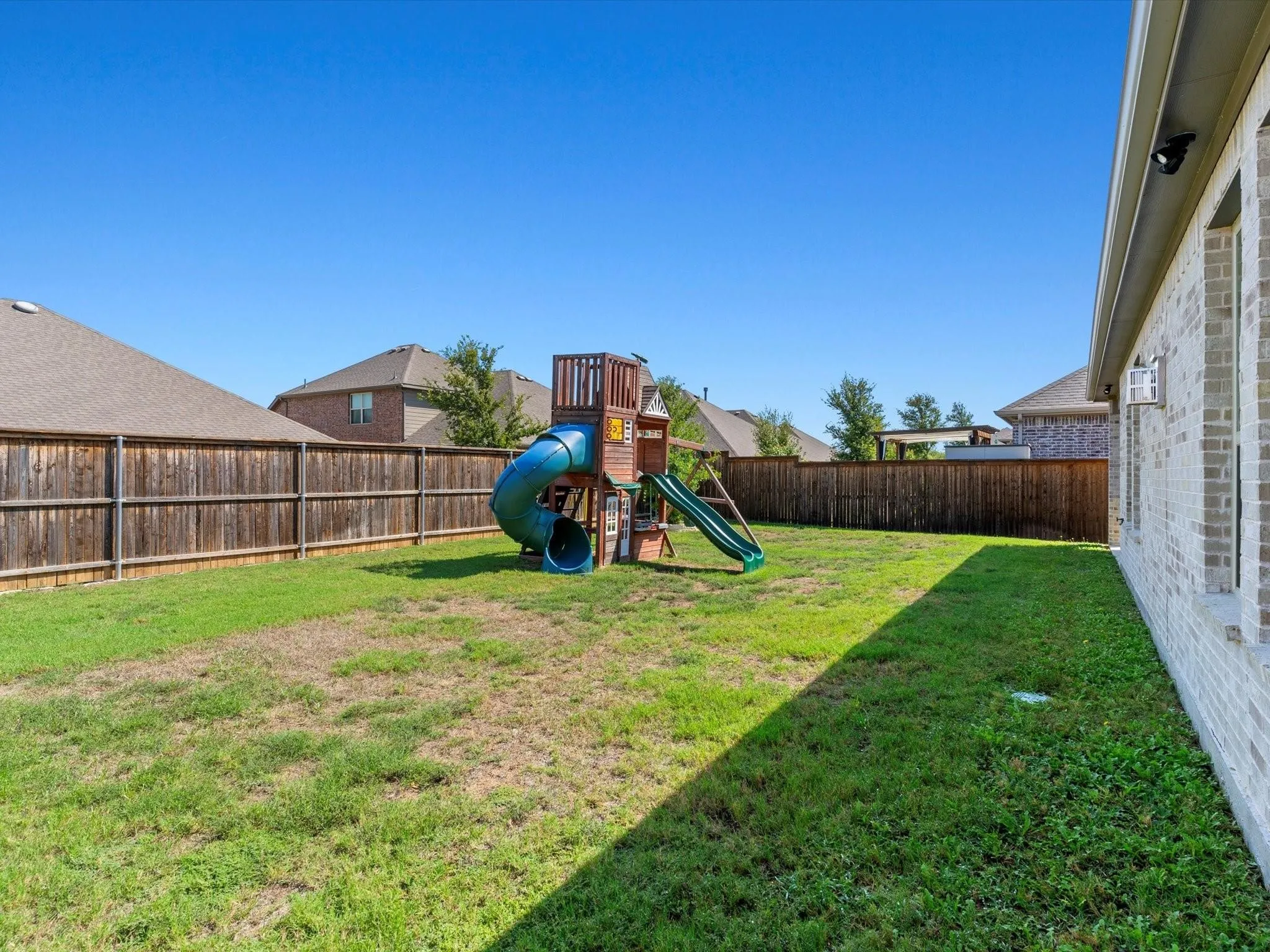 Spacious backyard featuring a wooden privacy fence, a brick exterior wall with windows, and a play structure