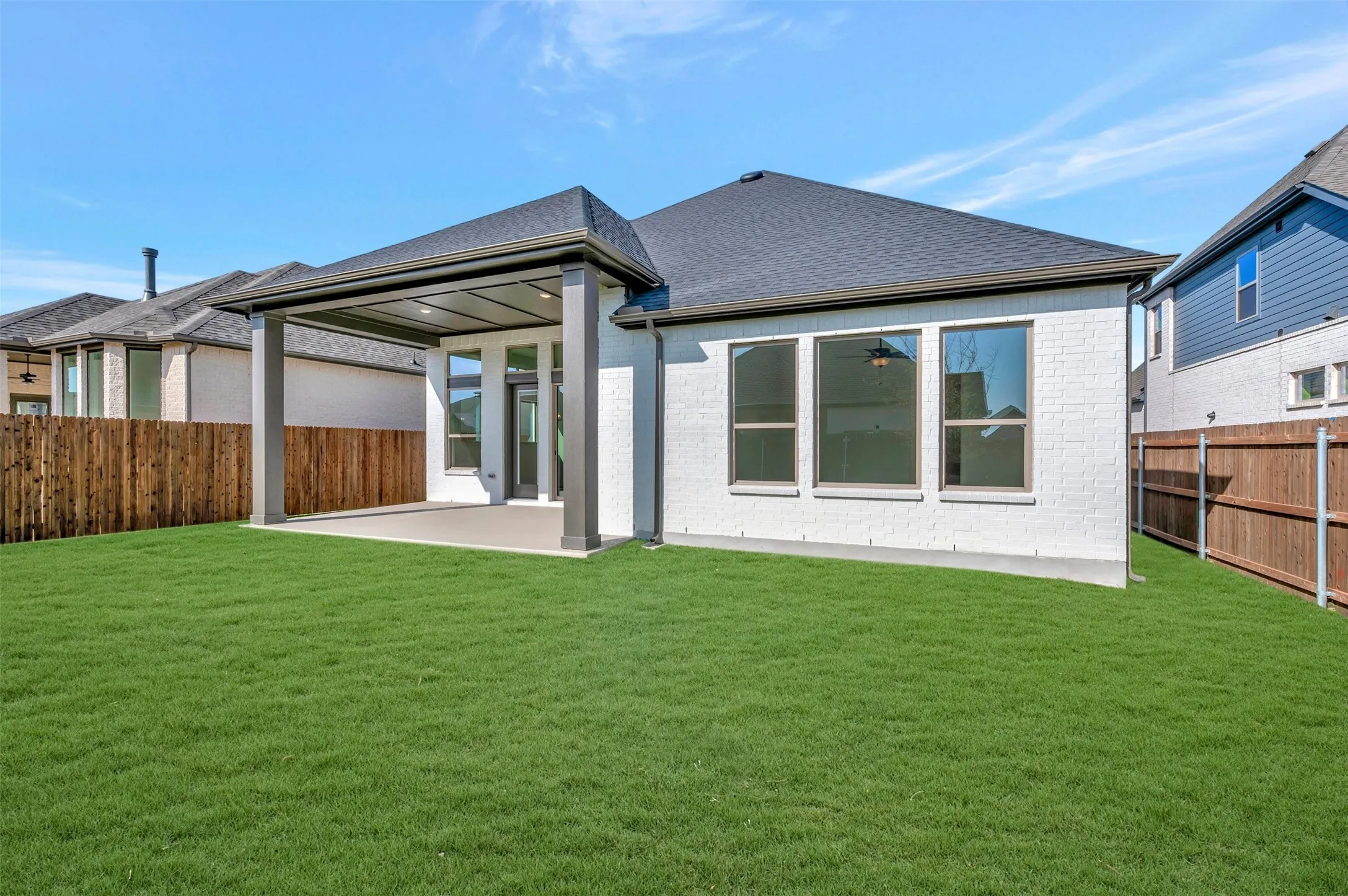Expansive green lawn with a covered patio, white brick exterior, dark grey roof, and a privacy fence