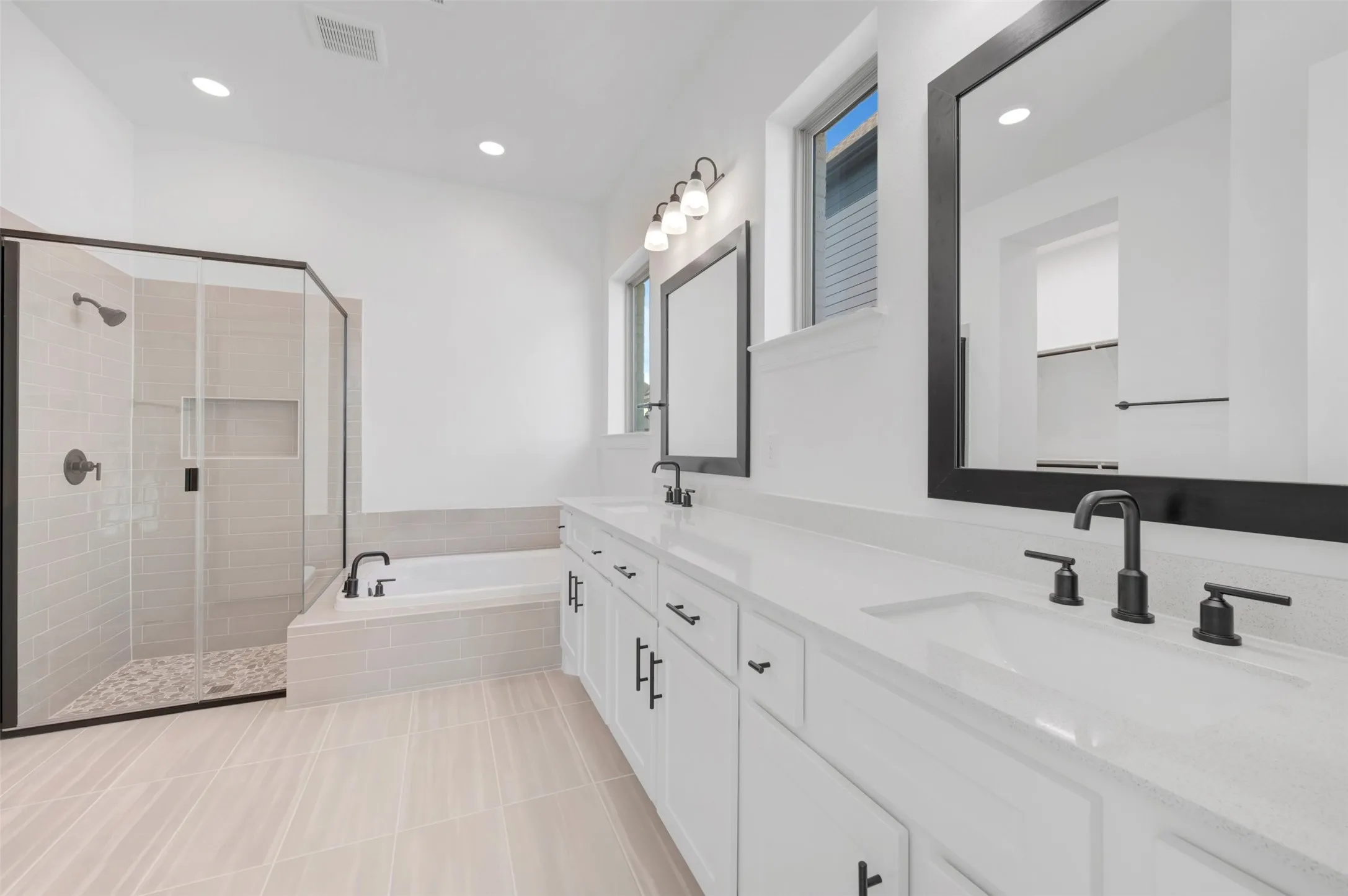 Contemporary bathroom featuring a spacious walk-in shower with pebble tile flooring and a built-in niche, a large soaking tub, a dual vanity with quartz countertops, framed mirrors, and matte black fixtures
