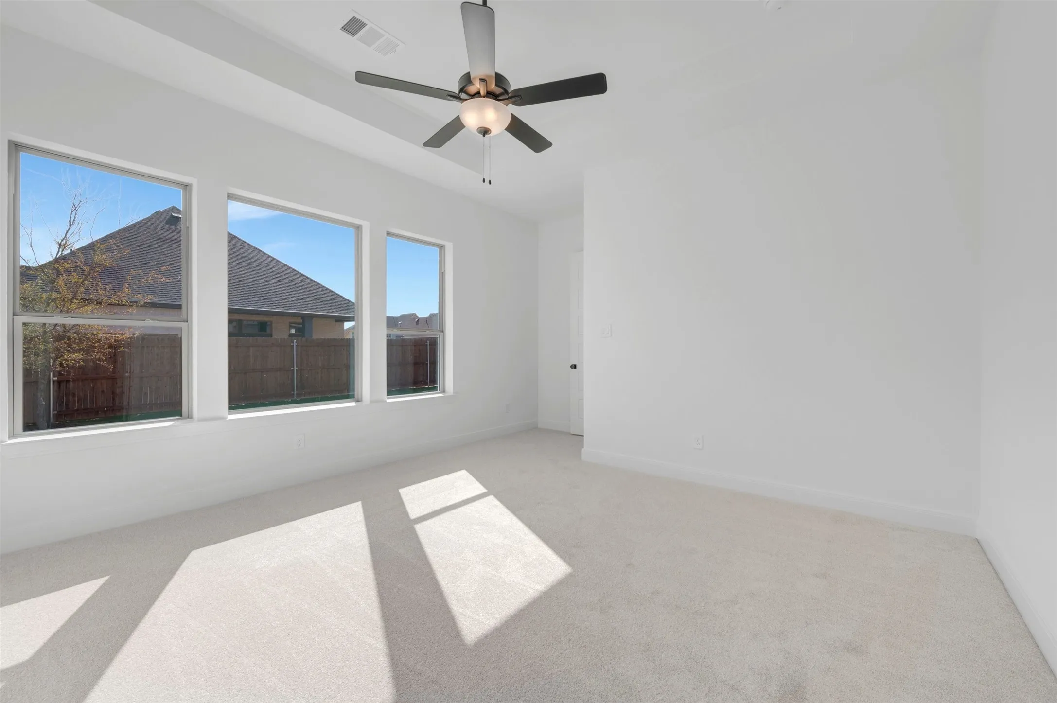 Spacious room featuring three large windows, light-colored carpet flooring, a ceiling fan with integrated lighting, and crisp white walls