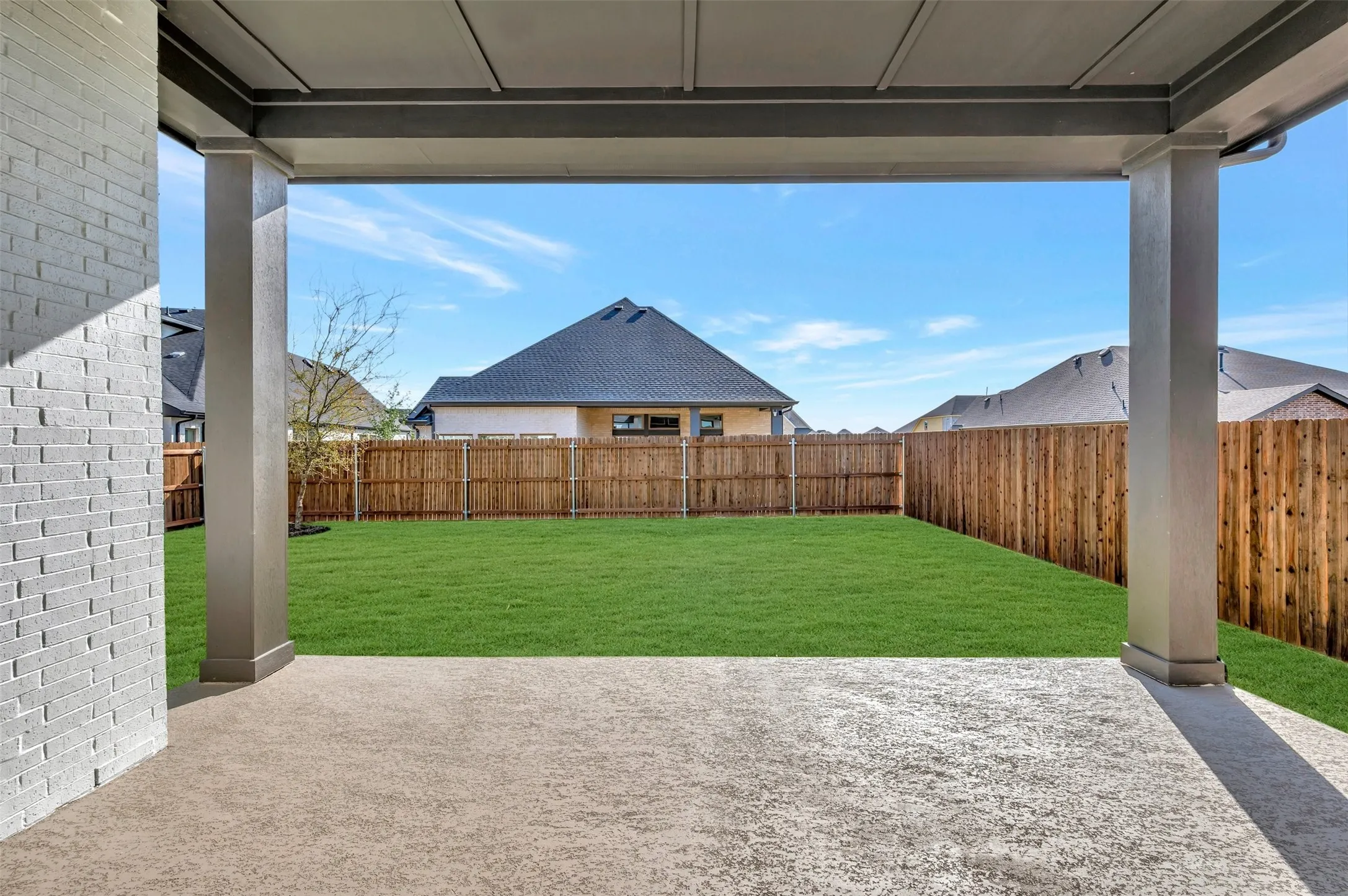 Covered patio featuring a concrete surface, supported by two columns, opening to a grass lawn, enclosed by a wood fence, and a brick exterior wall