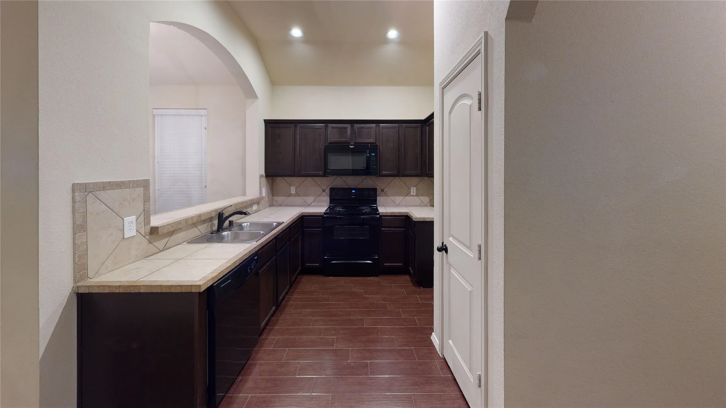 Kitchen featuring black appliances, tile countertops, tasteful backsplash, dark wood finish cabinetry, and wood tiled floors