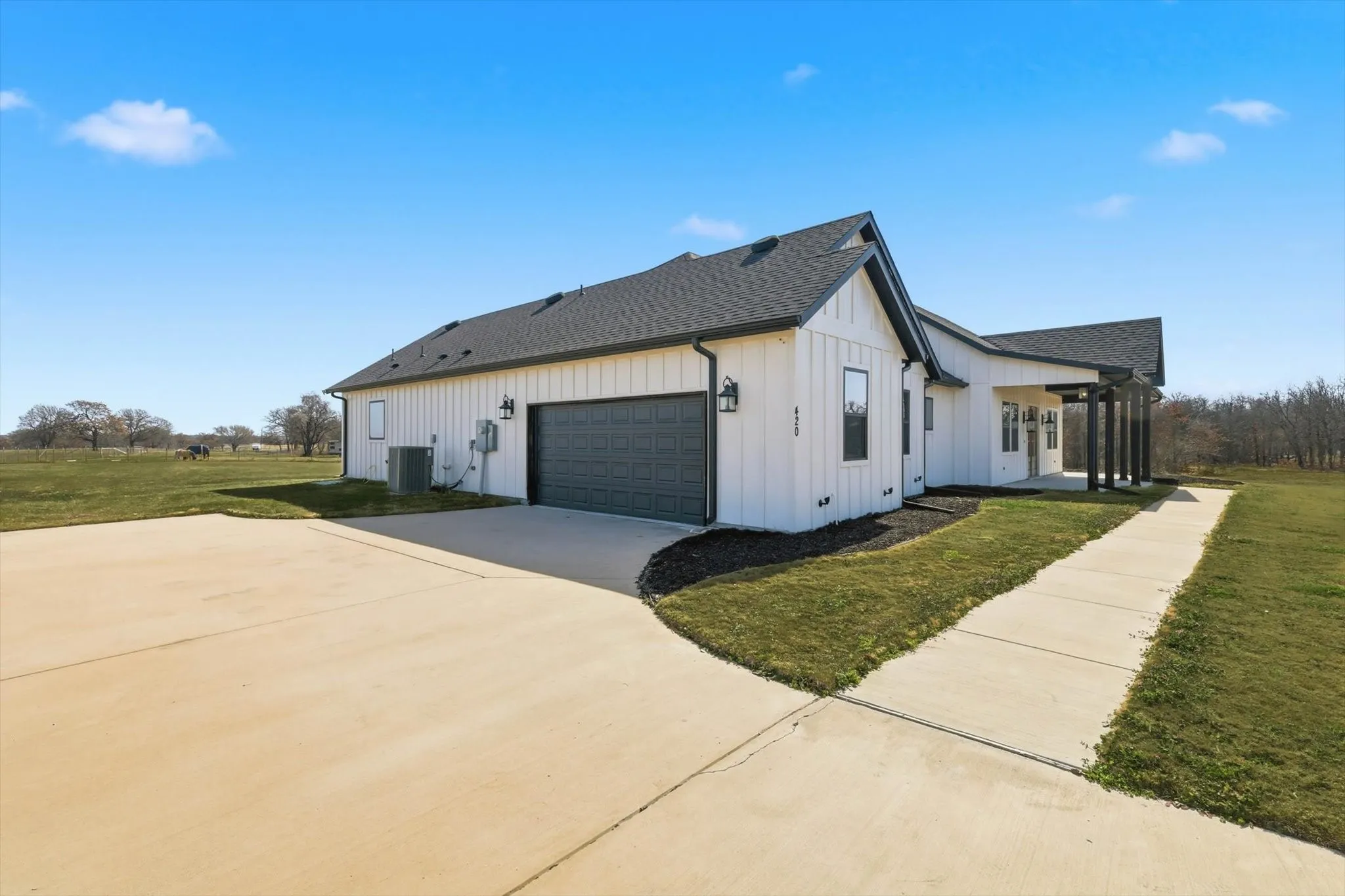 Wide-open arrival and plenty of parking. Oversized concrete drive leads to a clean modern farmhouse exterior with a two-car garage, crisp board-and-batten siding, and a covered walkway that pulls you right toward the front porch.