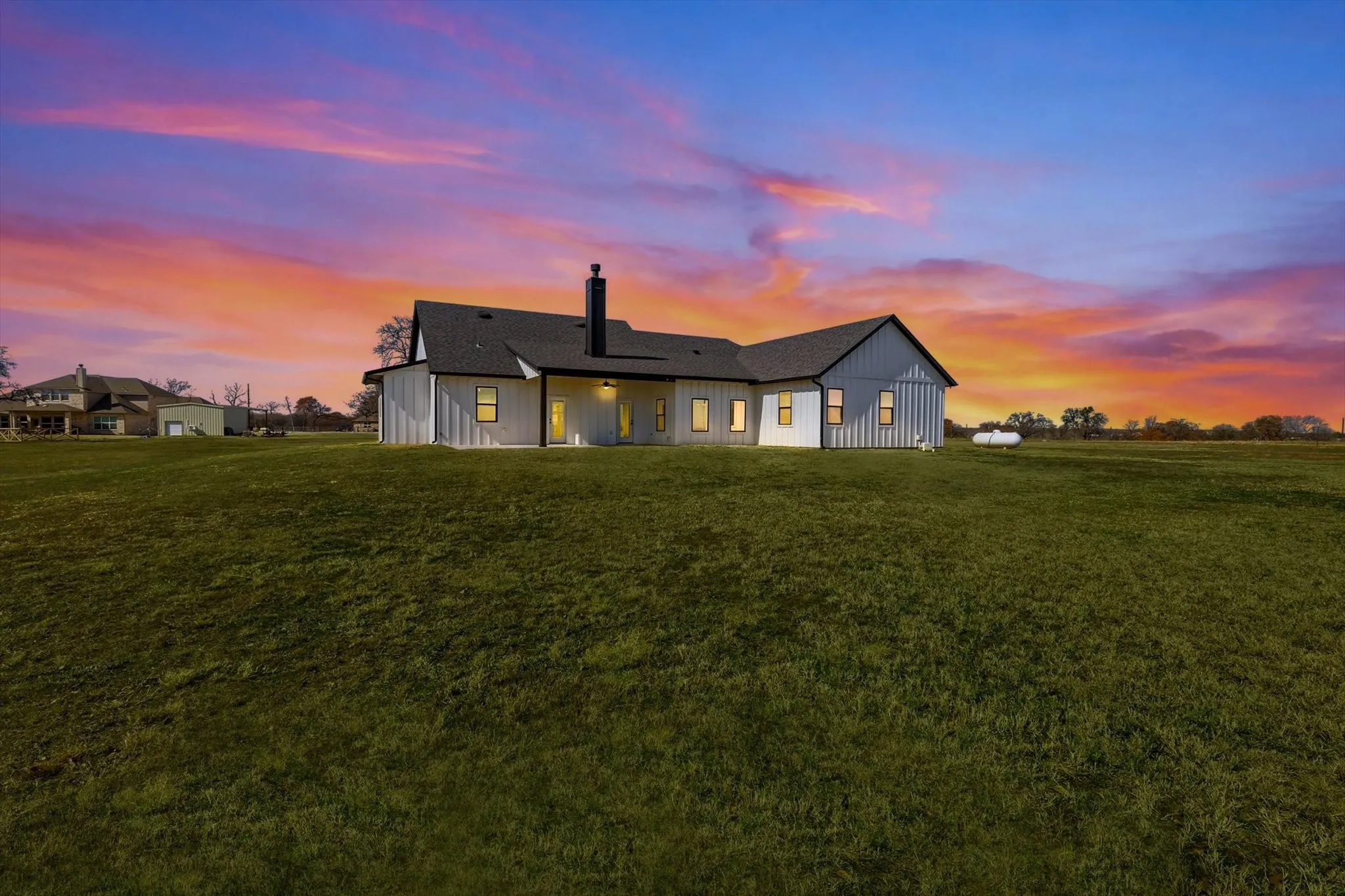 The back of the home offers a peaceful retreat with wide-open space, glowing interior light, and breathtaking sunset views stretching across the property.