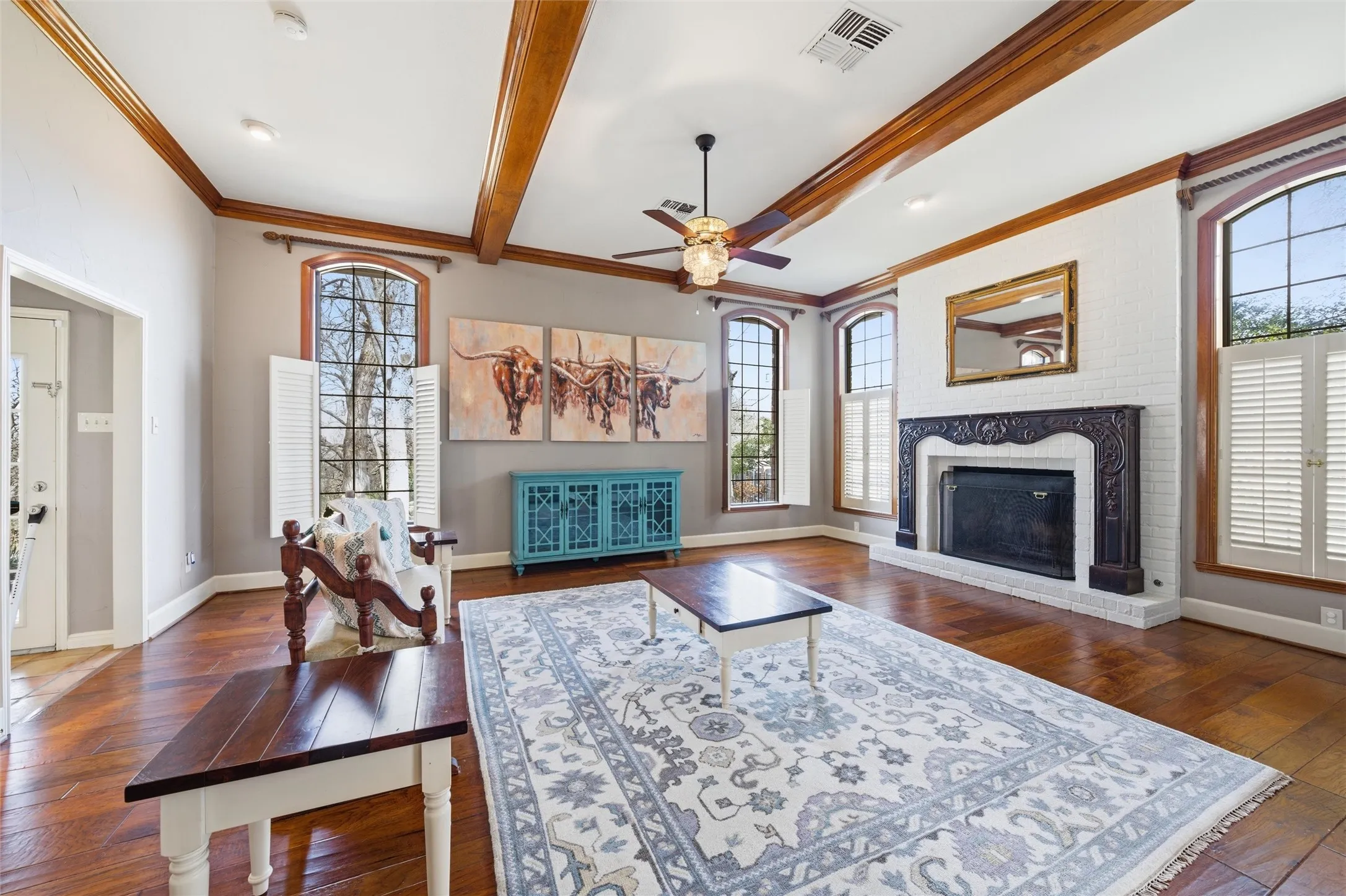 Living room featuring dark wood finished floors, a brick fireplace, ceiling fan, beamed ceiling, and ornamental molding