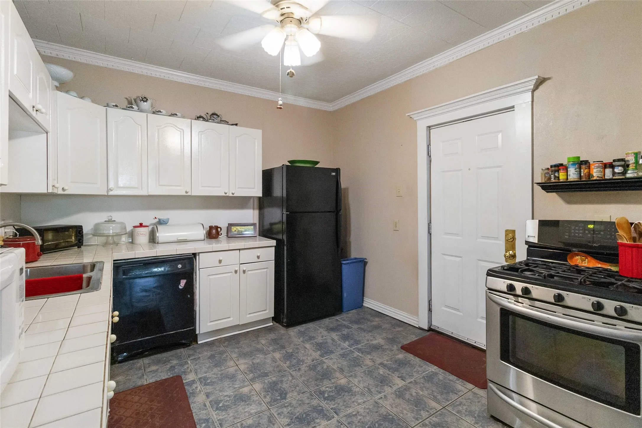 Kitchen featuring black appliances, white cabinets, a ceiling fan, tile countertops, and ornamental molding