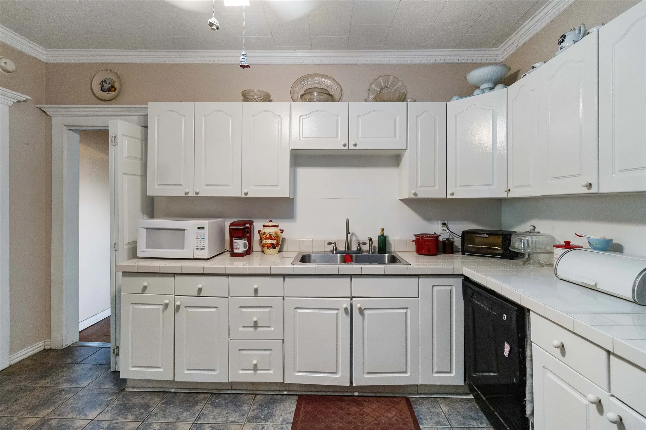 Kitchen featuring white microwave, white cabinets, ornamental molding, black dishwasher, and tile countertops