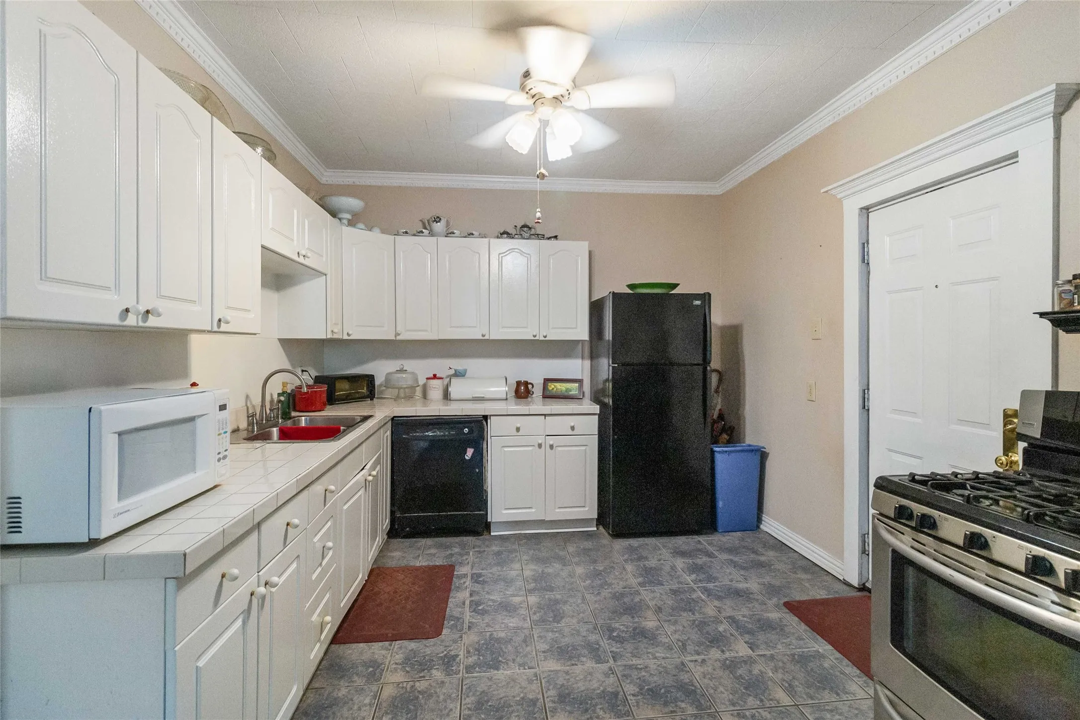 Kitchen featuring black appliances, a ceiling fan, white cabinets, tile countertops, and ornamental molding