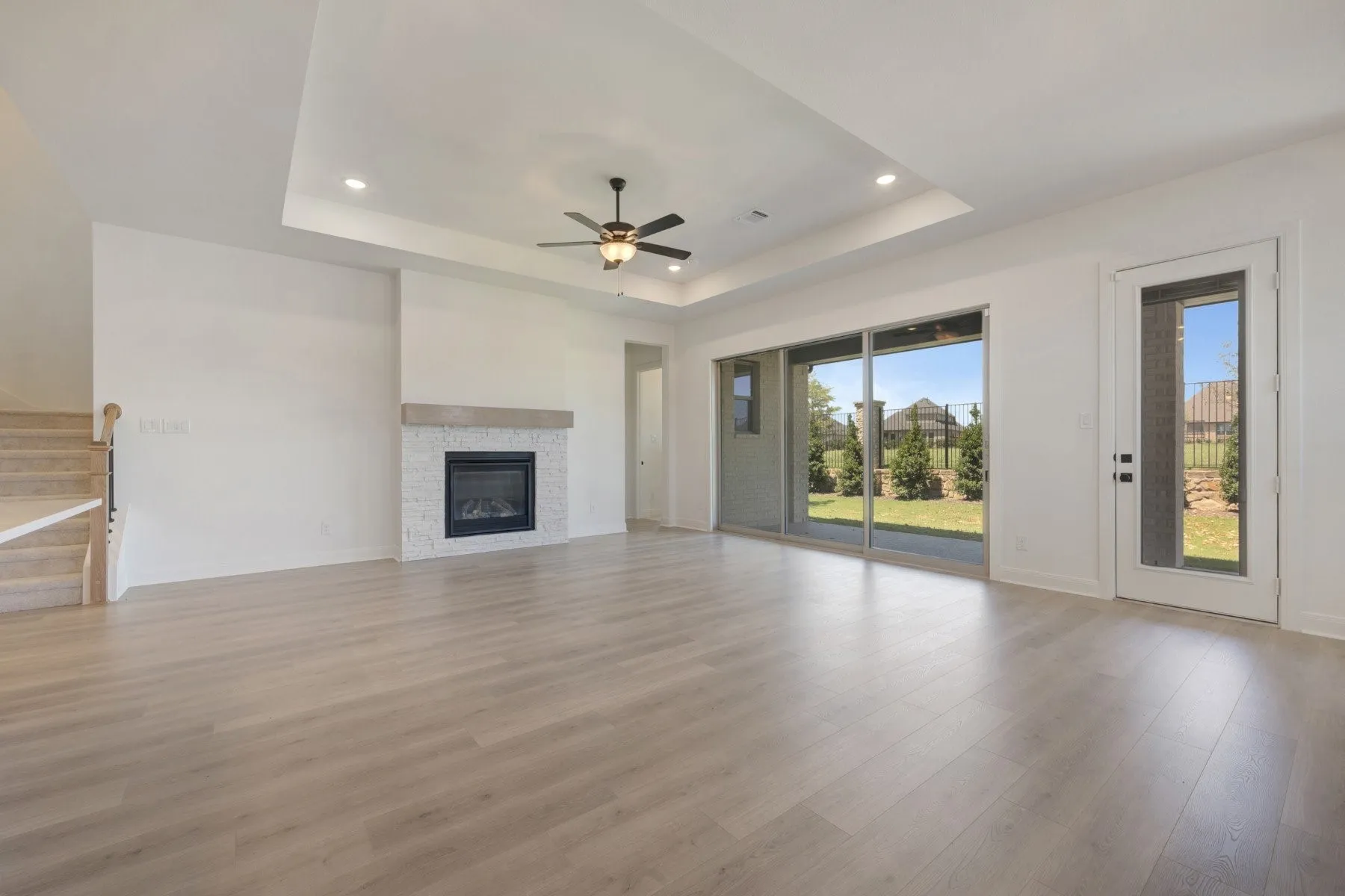 Spacious living area featuring wood-finish flooring, a contemporary fireplace with white stacked stone and a wood mantel, a tray ceiling with recessed lighting, and a ceiling fan