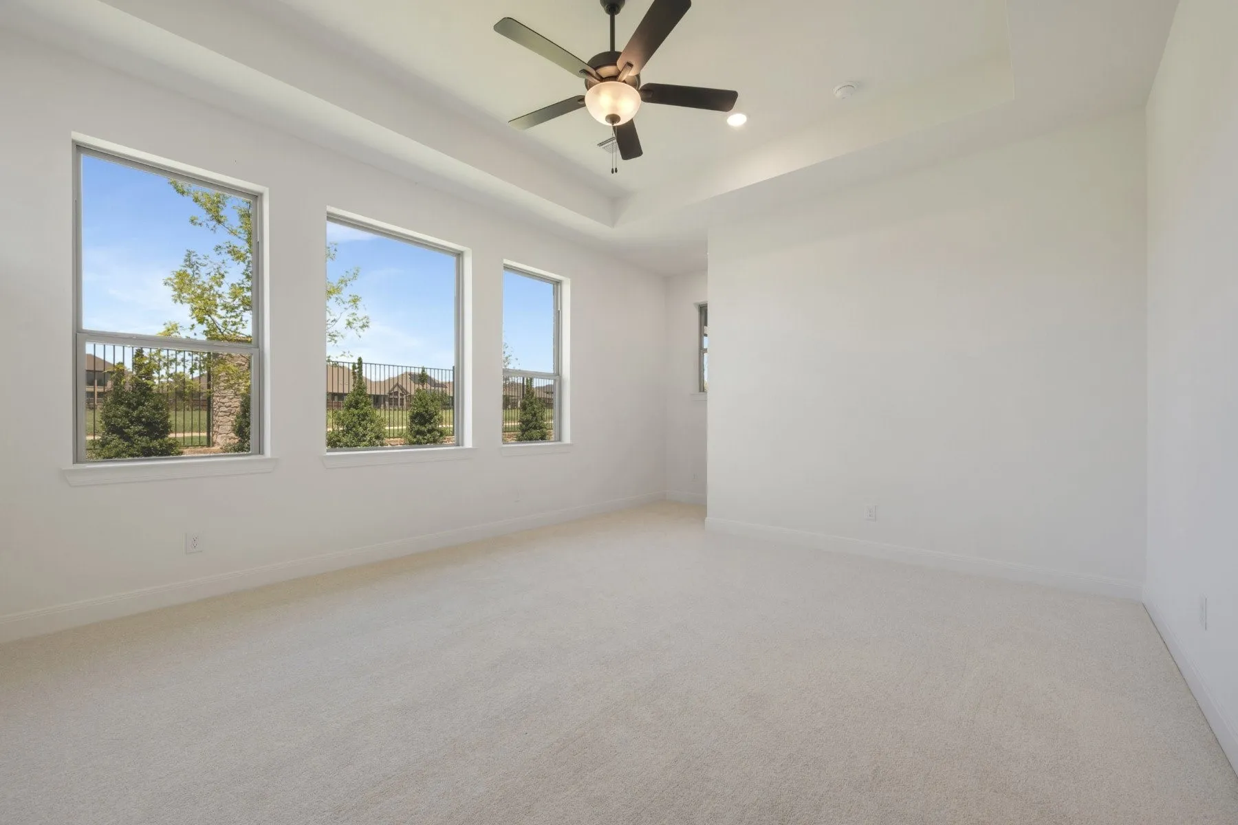 Bright room featuring a tray ceiling with a built-in ceiling fan, three large windows, and neutral carpet flooring