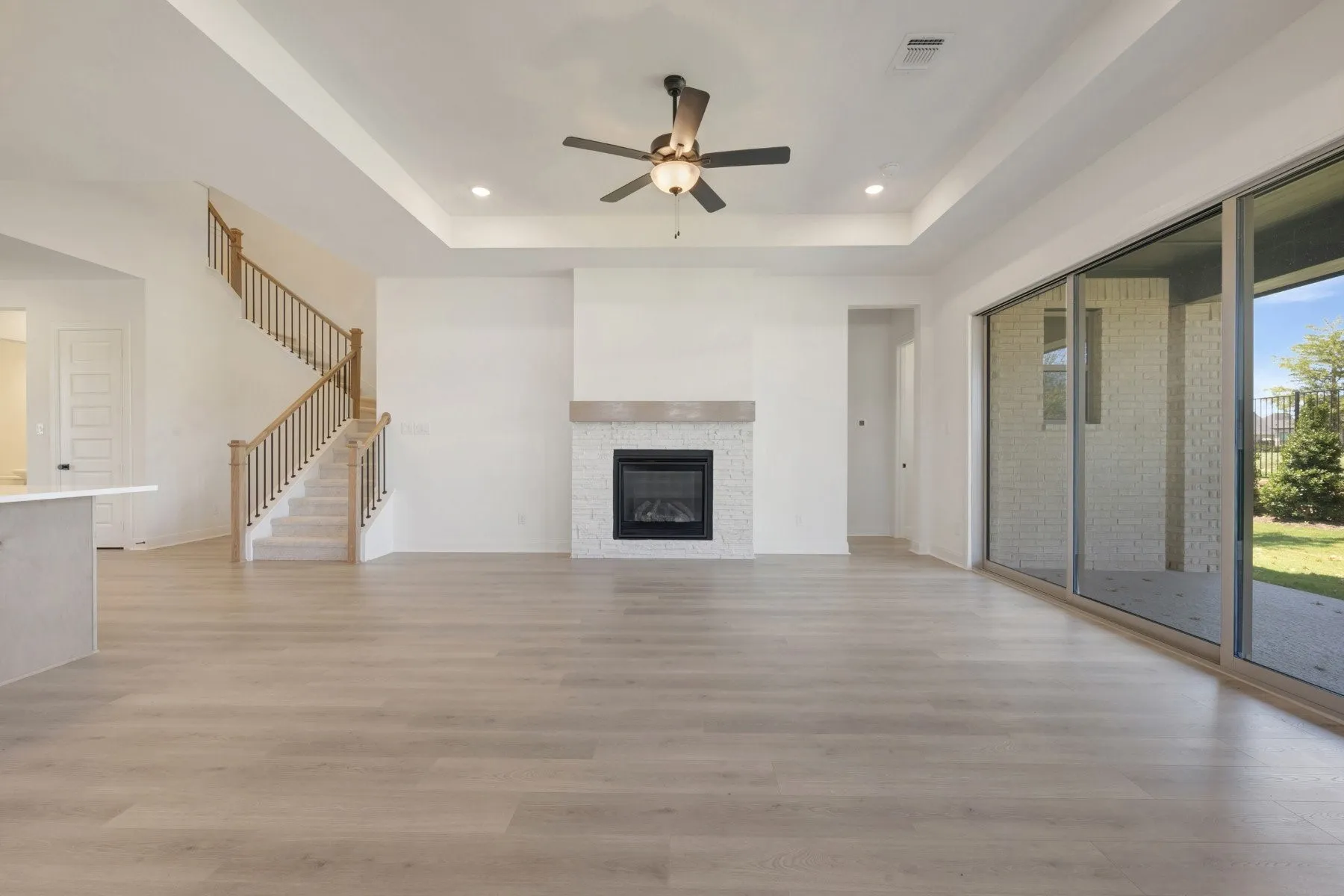 Spacious living area featuring wood-finish flooring, a contemporary fireplace with stone tile surround and mantel, and a tray ceiling with recessed lighting and ceiling fan