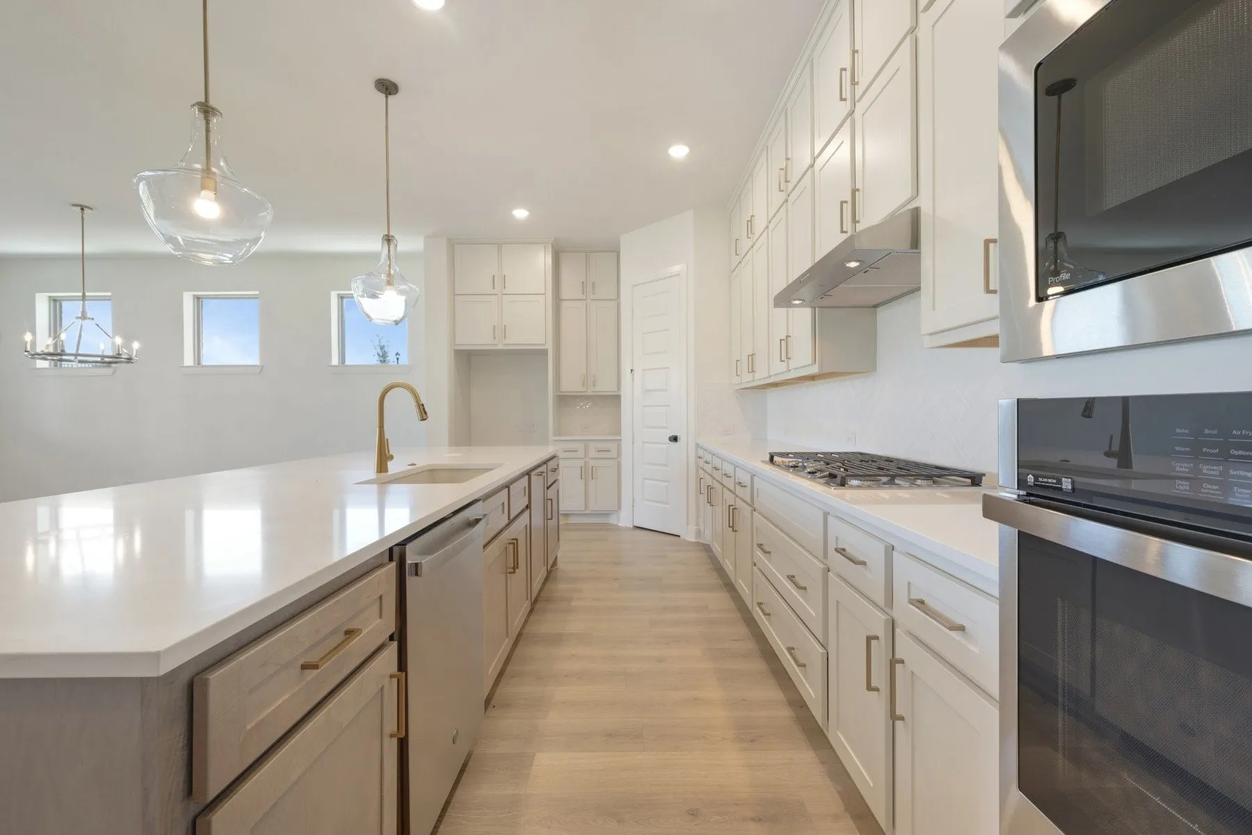 Bright kitchen featuring a large island with sink, white shaker-style cabinetry, stainless steel appliances, light wood-finish flooring, and pendant lighting