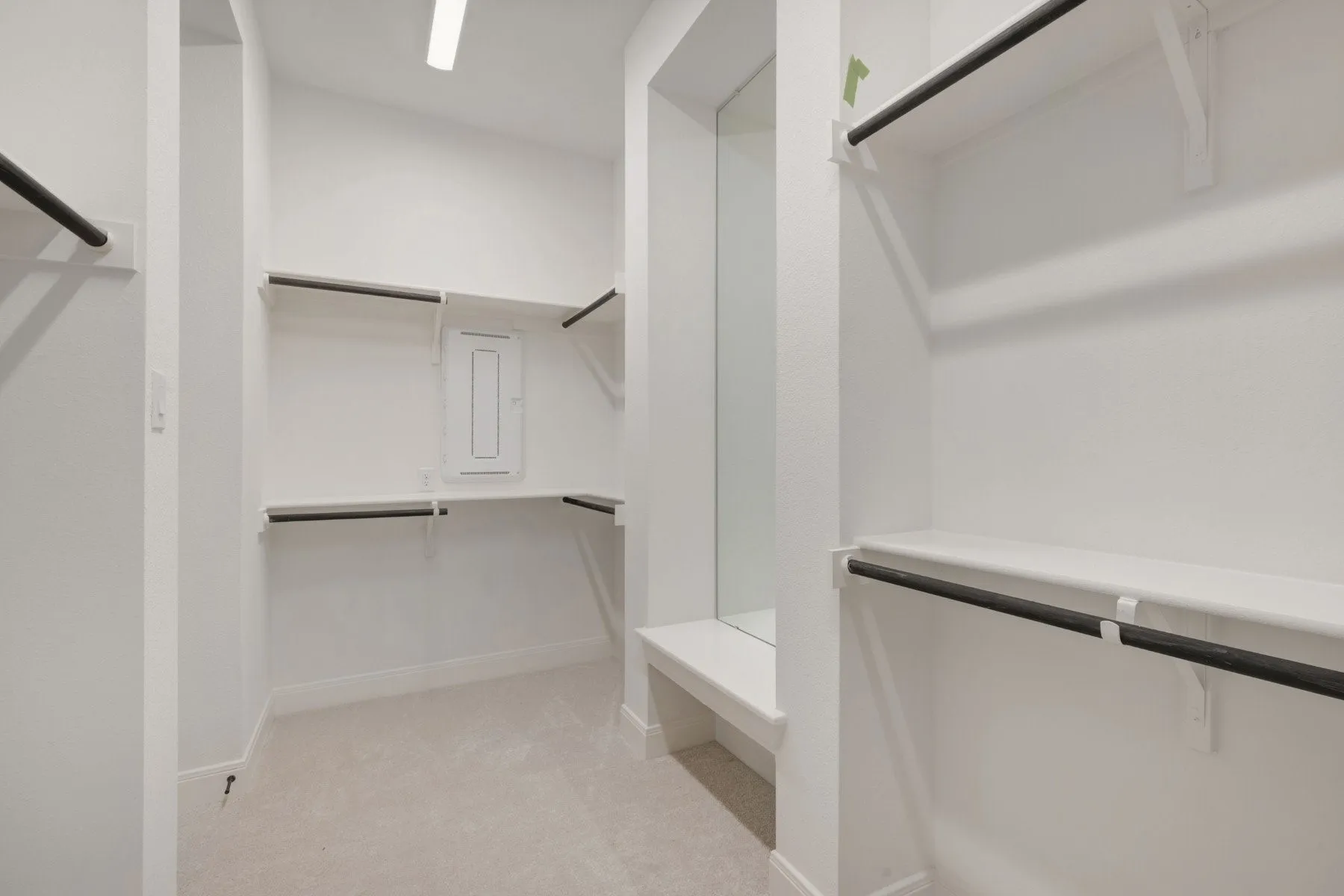Spacious walk-in closet featuring white shelving, matte black hanging rods, light-toned carpeting, and an integrated dressing bench with a full-length mirror