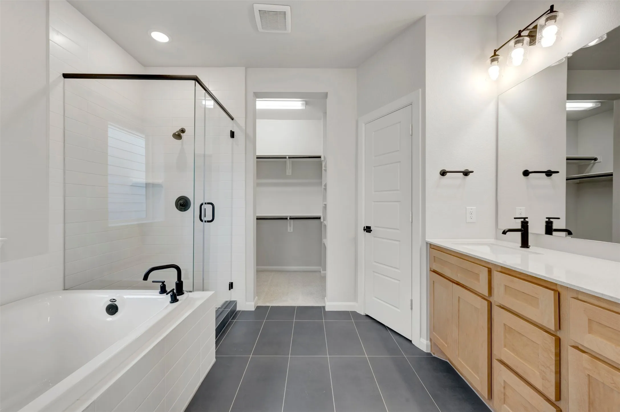 Spacious bathroom featuring a built-in tub, separate glass-enclosed shower, dual-sink vanity with light wood cabinetry, and extensive built-in closet shelving