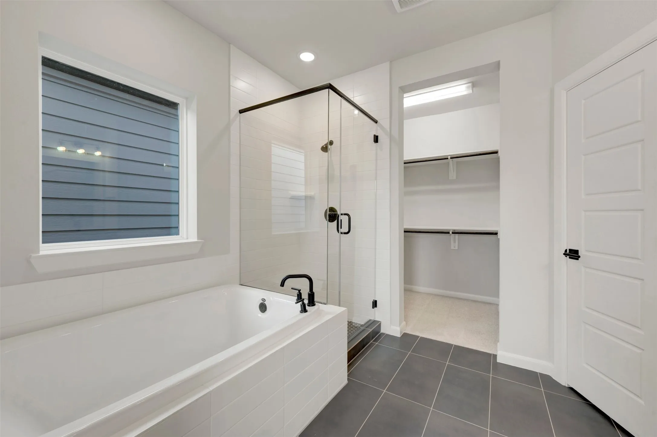 Deep soaking tub with matte black fixtures, glass-enclosed shower, dark tile flooring, and a walk-in closet with built-in shelving