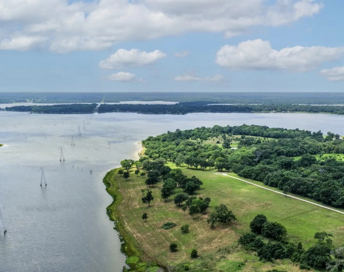 Expansive waterfront property featuring a natural shoreline, mature trees, open green spaces, and a gravel road traversing the landscape
