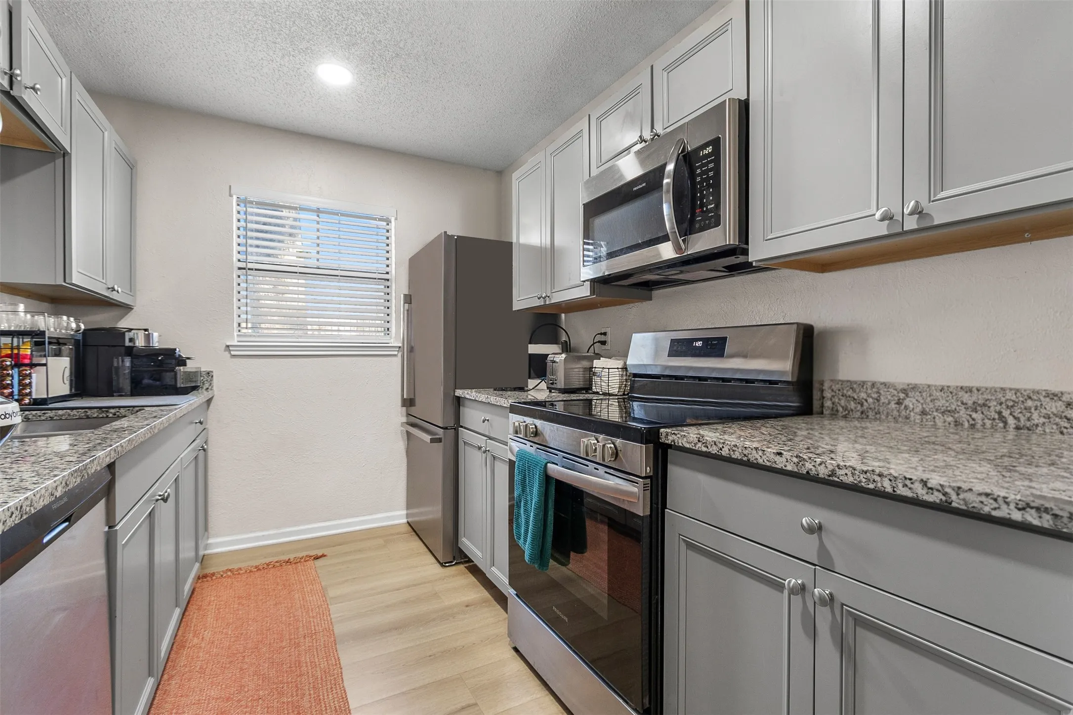 Kitchen with gray cabinetry, stainless steel appliances, a textured ceiling, light stone counters, and light wood-style floors