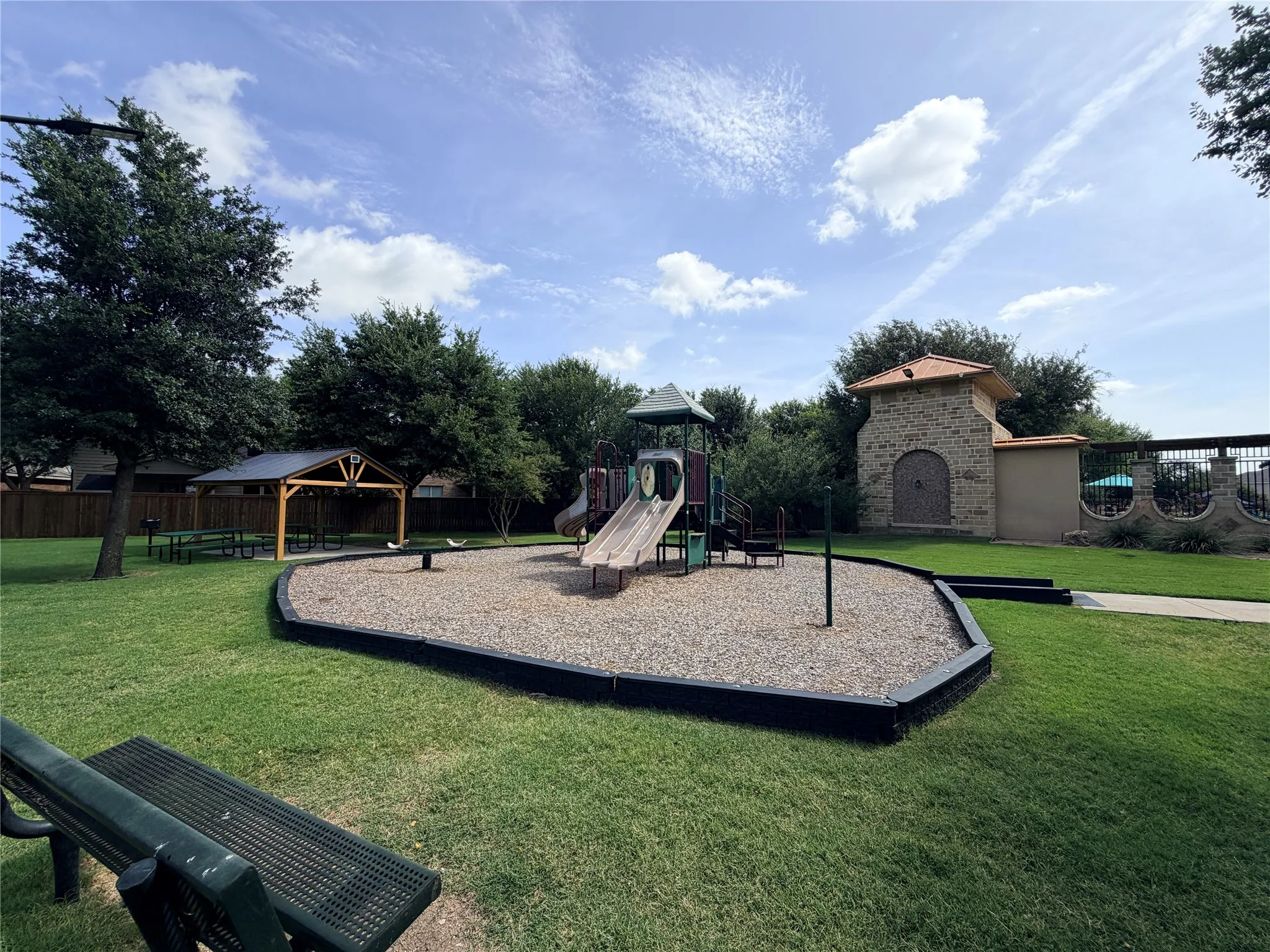 Community park featuring a dedicated playground area with play structure and slides, an adjacent covered picnic area, and mature trees providing natural shade