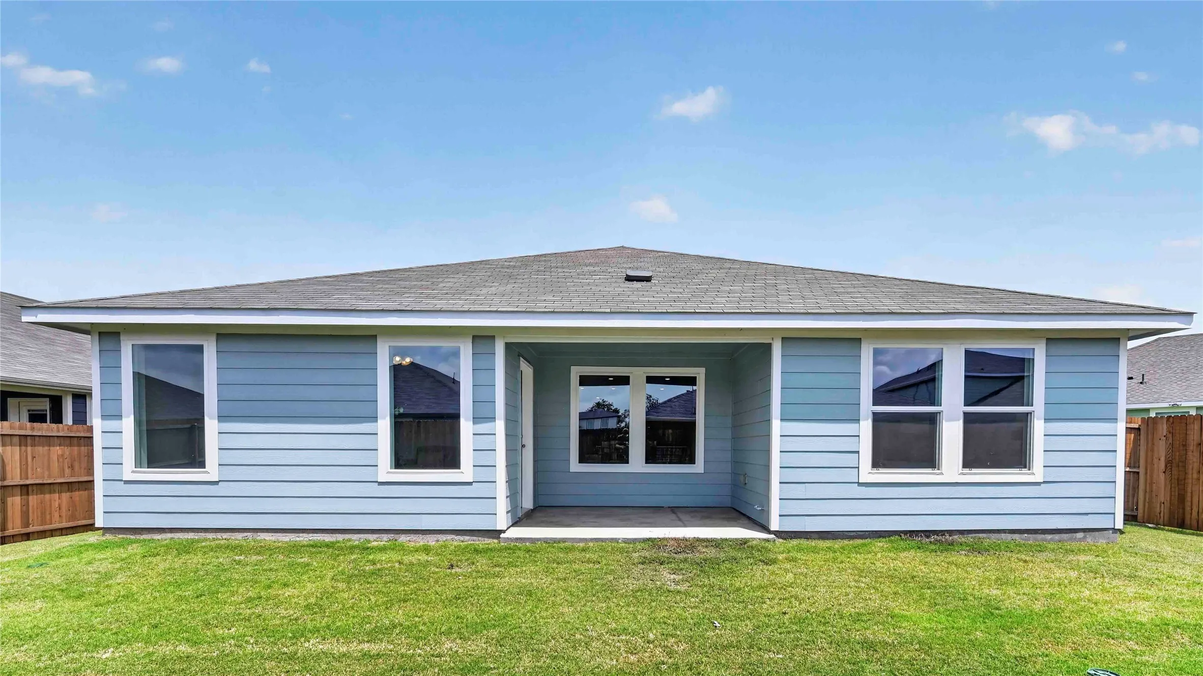 Rear exterior featuring light blue siding, a covered patio with concrete flooring, multiple windows with white trim, and a shingled roof