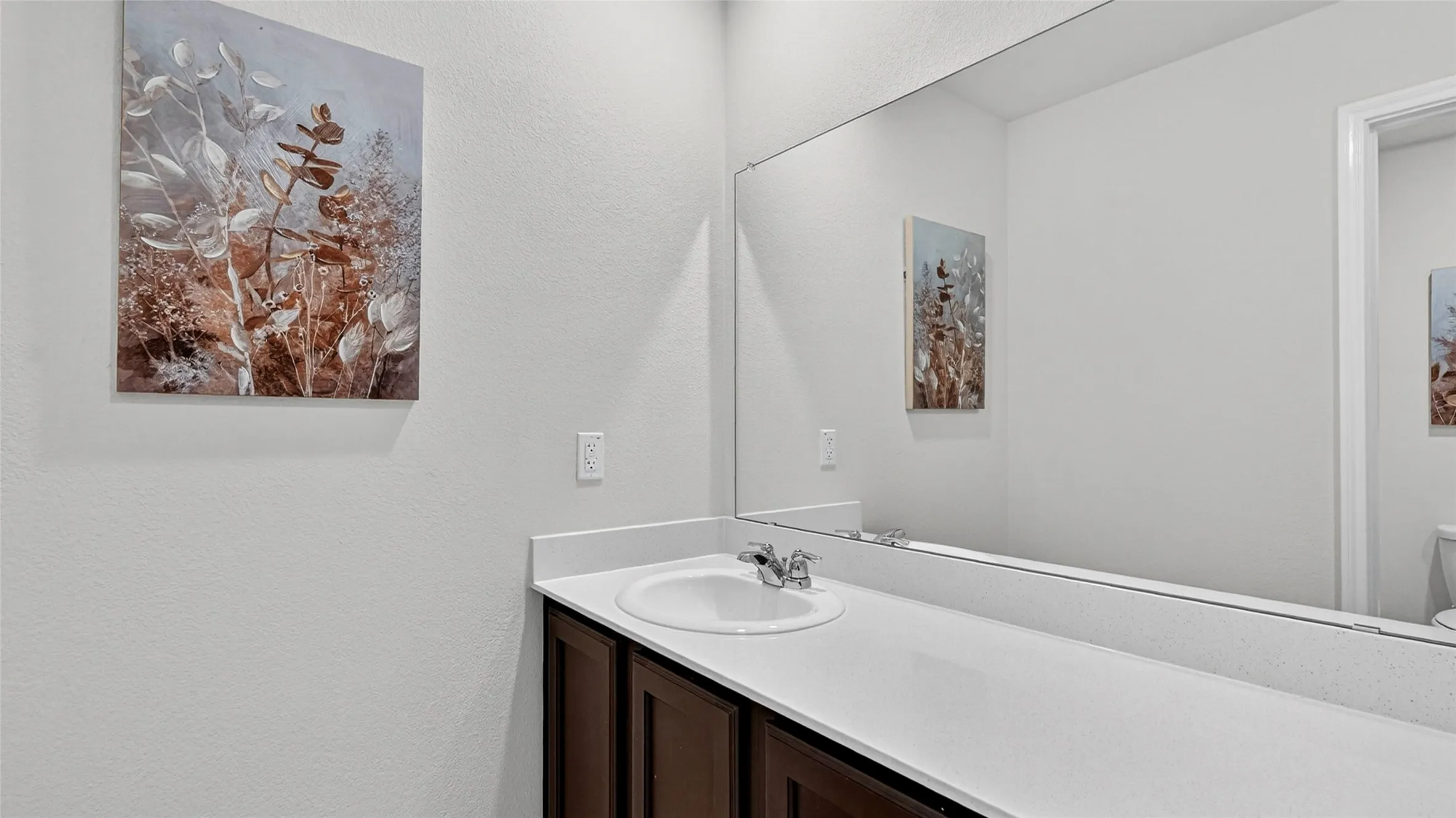 Bathroom vanity featuring a built-in sink with a chrome faucet, solid surface countertop, dark wood-finish cabinetry, and a full-width mirror