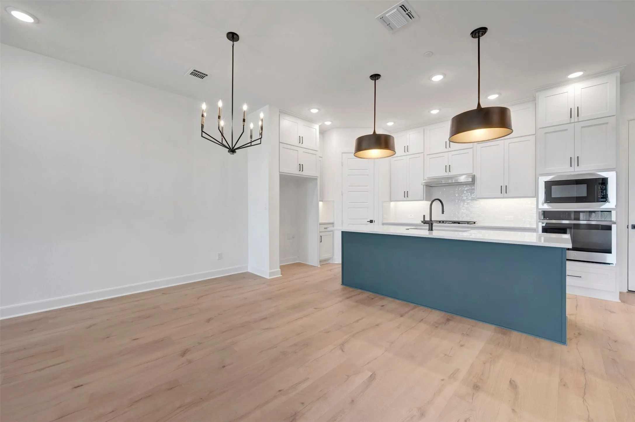 Open-concept kitchen and dining area featuring light wood-finish flooring, a contemporary black chandelier, a central island with a blue base and white countertop, white shaker cabinetry, and stainless steel appliances
