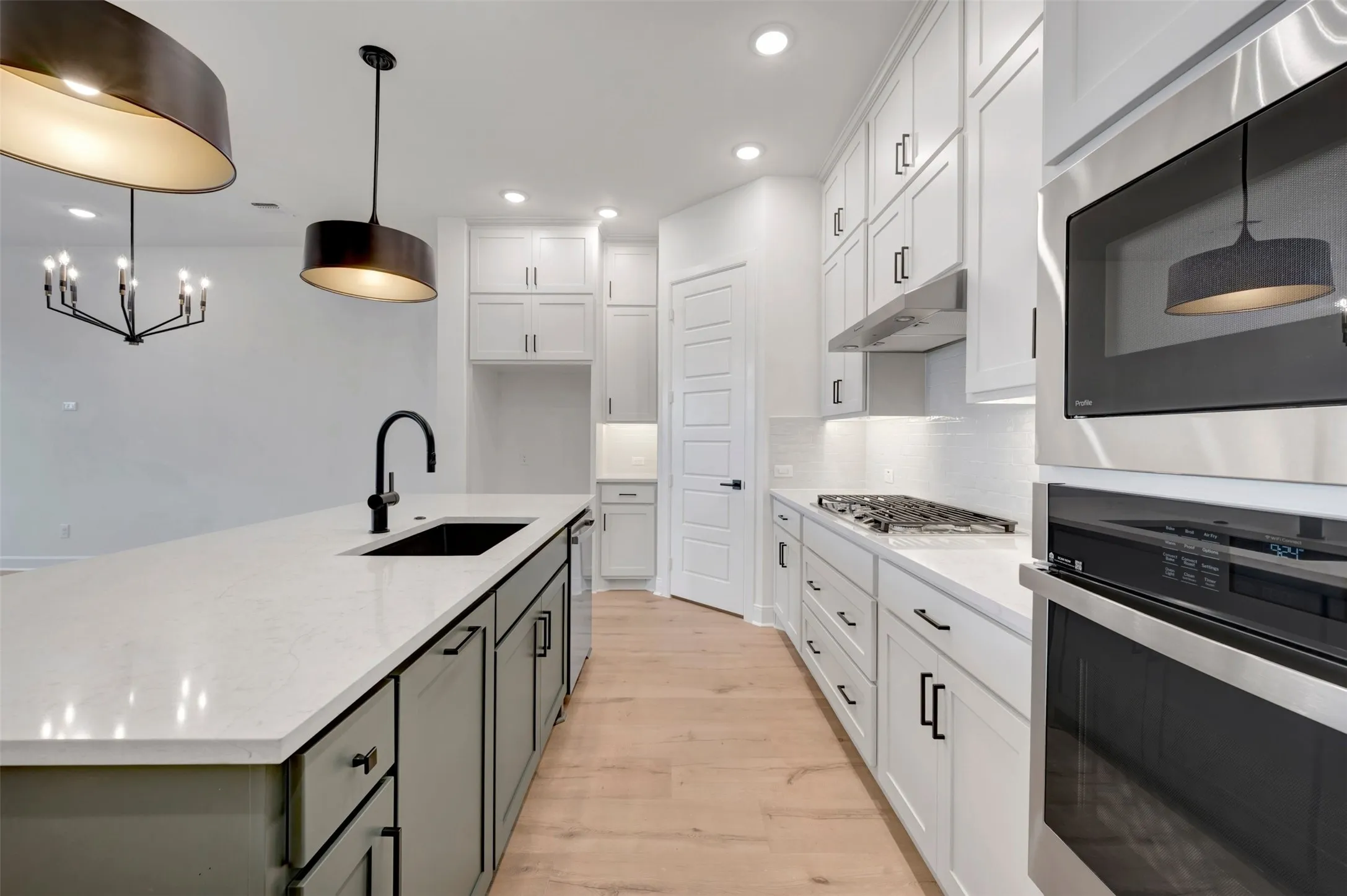 Contemporary kitchen featuring white shaker cabinetry, stainless steel appliances, a center island with a black sink and matte black faucet, light wood-finish flooring, and pendant lighting fixtures