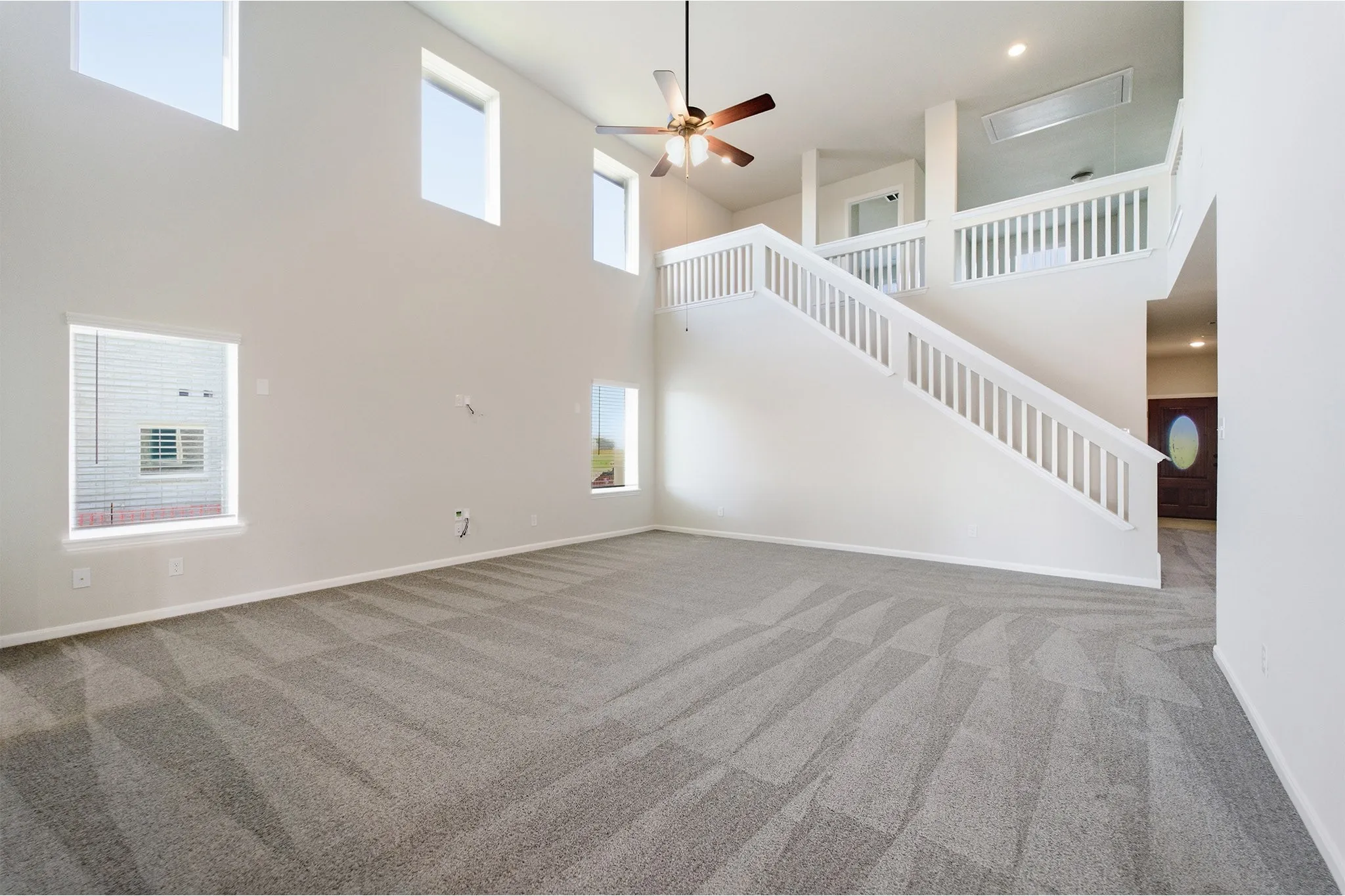 Two-story living area featuring a vaulted ceiling with a ceiling fan, light gray carpet, multiple windows, and a white staircase with balusters leading to an upper landing