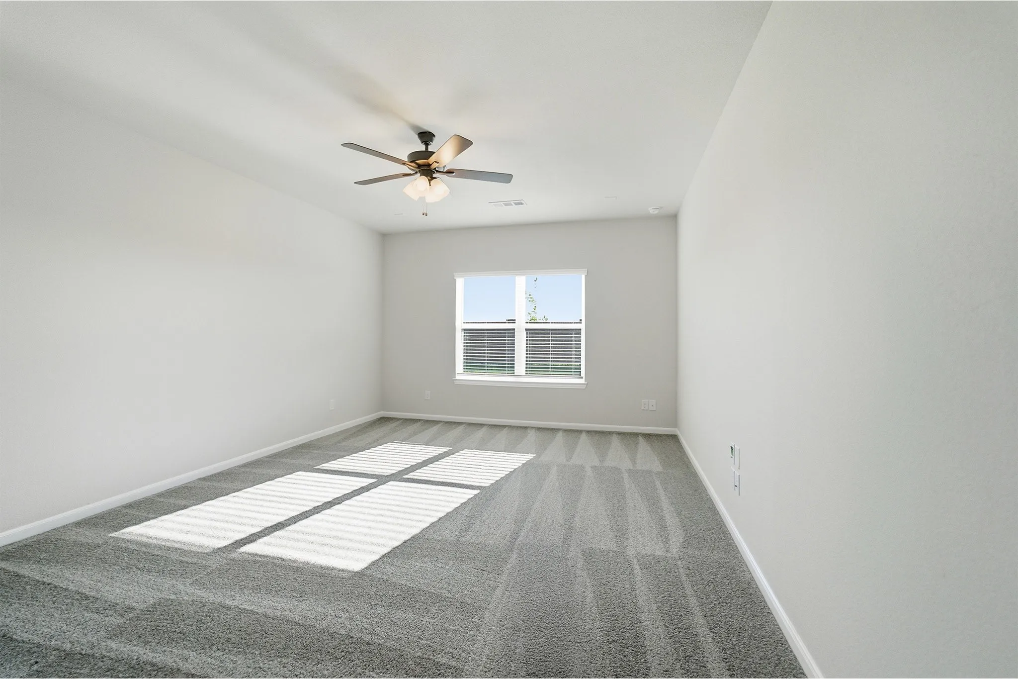 Spacious room featuring neutral painted walls, textured gray carpeting, a multi-pane window with horizontal blinds, and a ceiling fan with integrated lighting