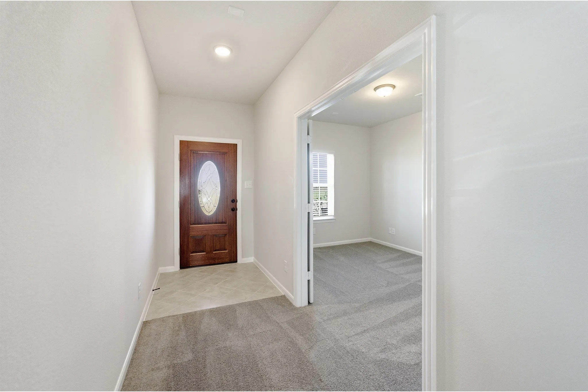 Entryway featuring a rich wood-finish front door with an oval glass insert, recessed lighting, and tile flooring transitioning to soft gray carpeting