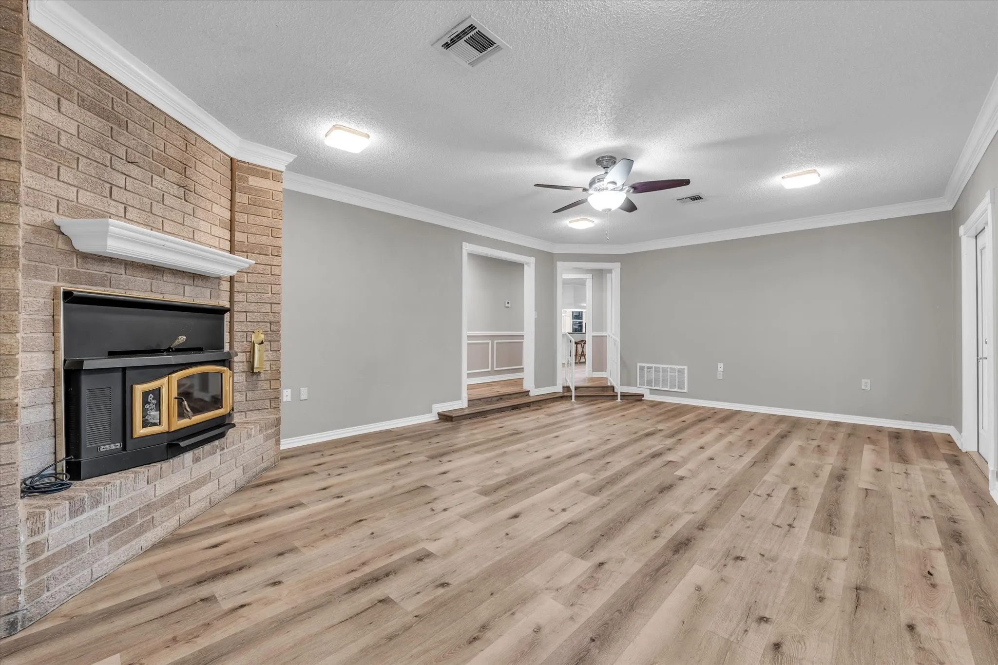 Unfurnished living room with a brick fireplace, light wood-style floors, a ceiling fan, a textured ceiling, and crown molding