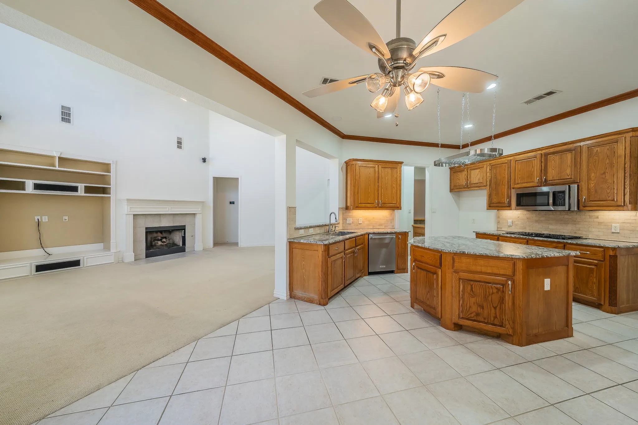 Open-concept living and kitchen space featuring wood cabinetry with stainless steel appliances, tile flooring throughout, a center island with seating, vaulted ceiling with ceiling fan, and a fireplace in the adjoining living area