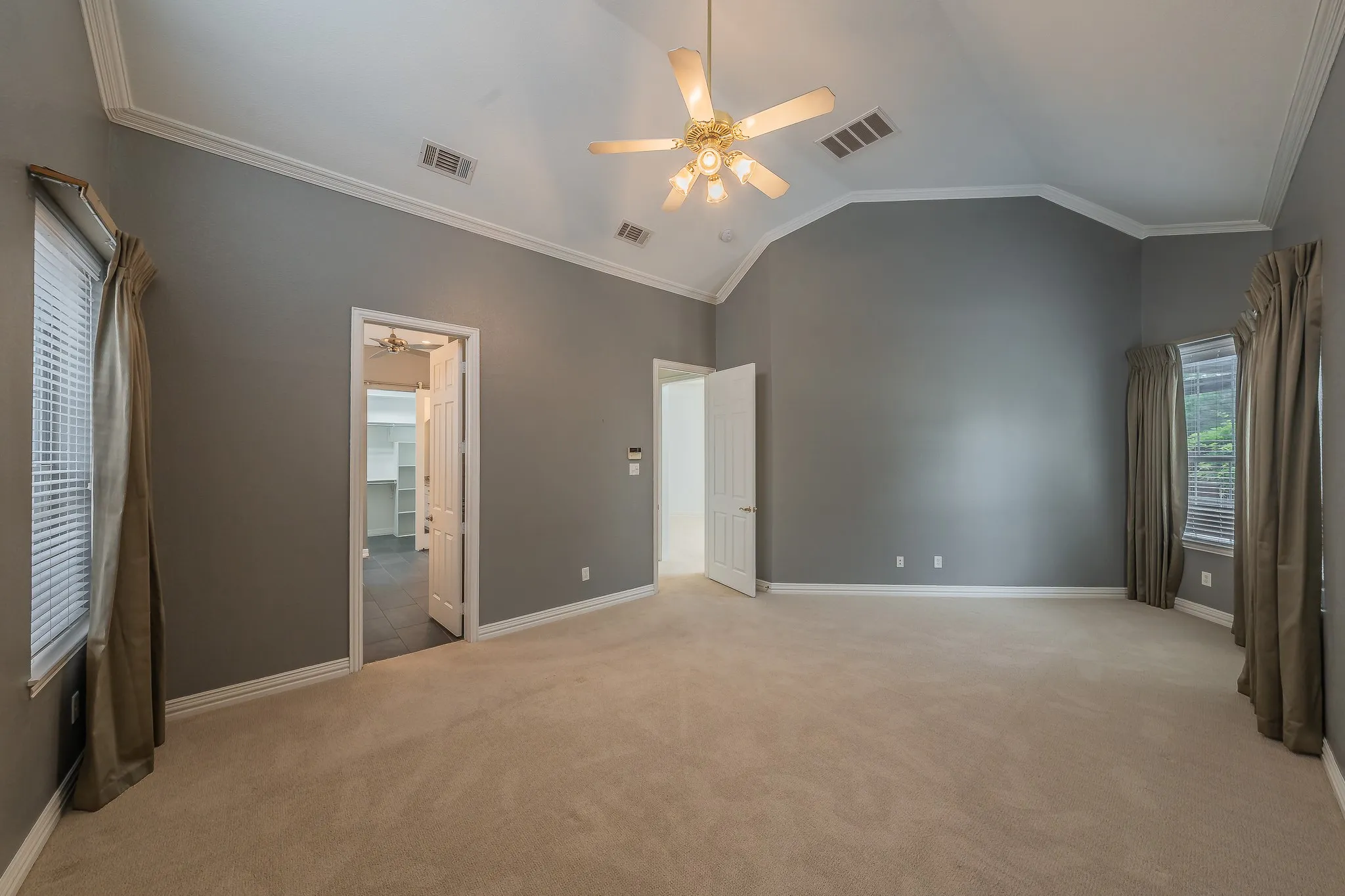 Spacious primary bedroom featuring vaulted ceilings, crown molding, a ceiling fan with light fixture, neutral carpeting, and multiple windows with blinds also with sitting area.