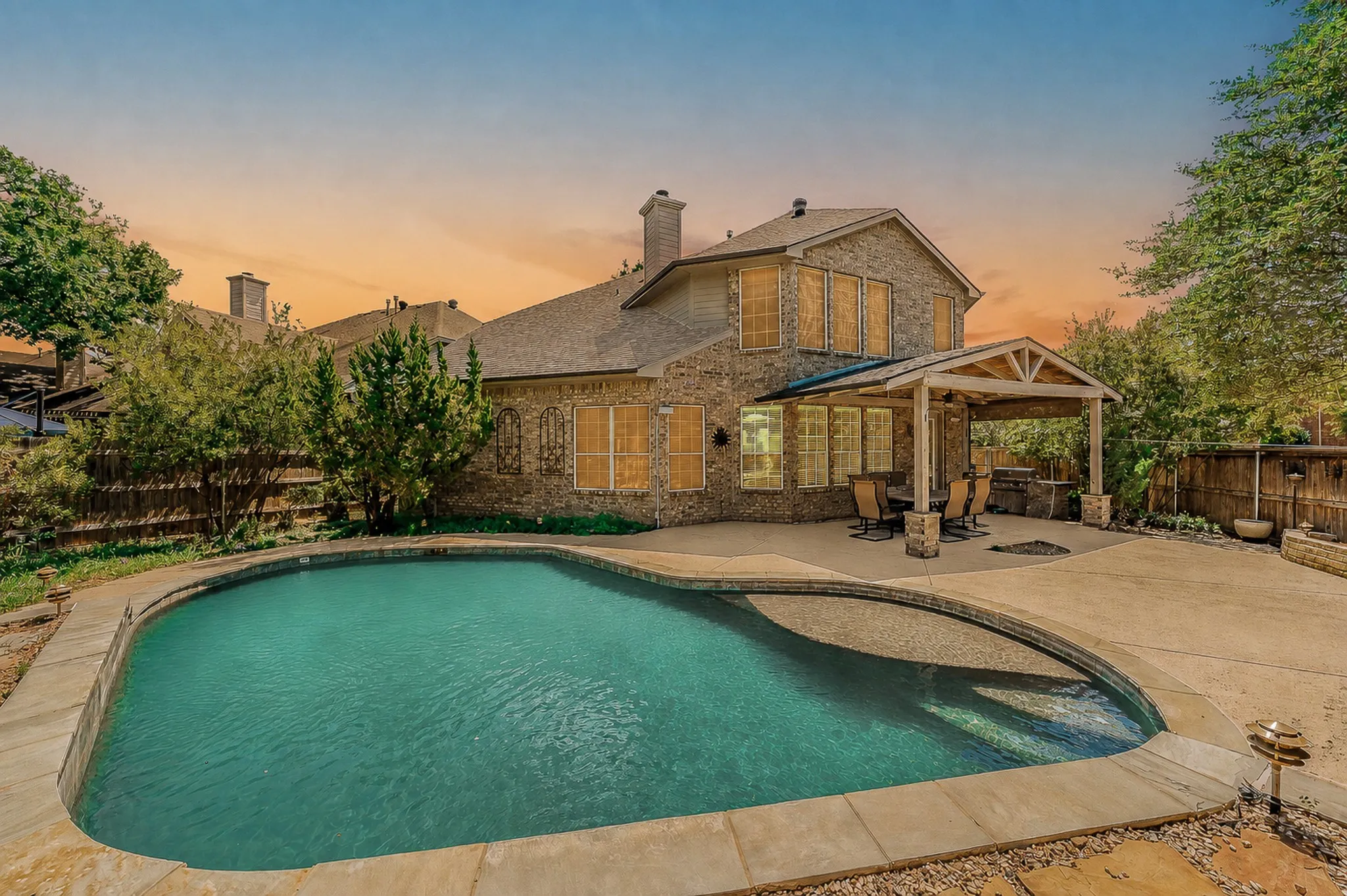 In-ground swimming pool with stone coping, expansive patio, covered outdoor living area, brick exterior, and wood fencing with gorgeous sunset view.
