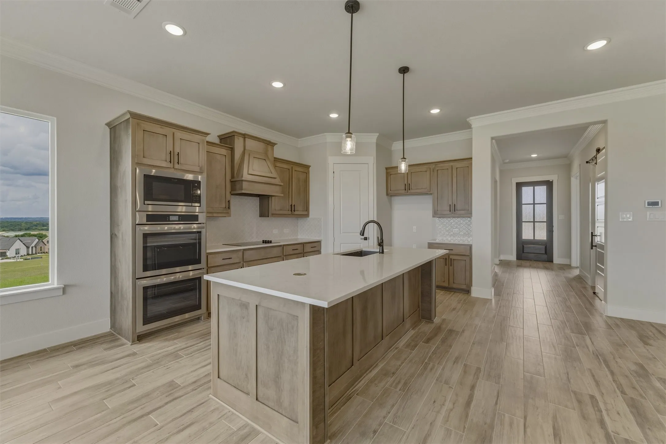 Spacious kitchen featuring a central island with sink, light wood-finish cabinetry, built-in stainless steel double ovens, and wood-finish flooring