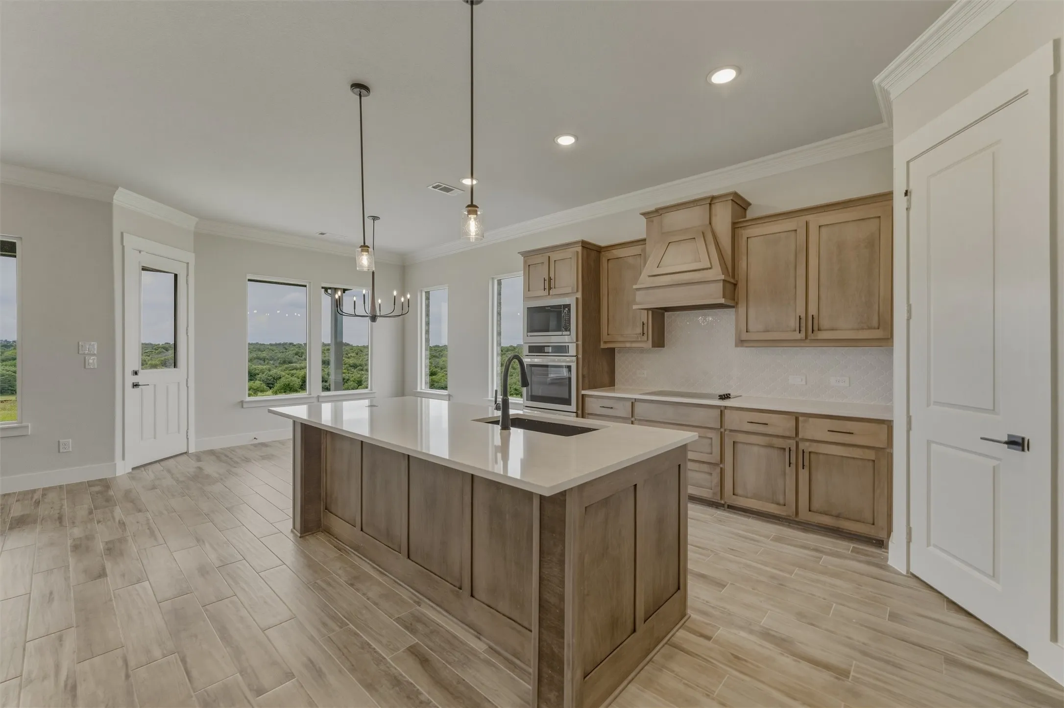 Gourmet kitchen featuring a large island with sink, integrated double ovens, wood-finish cabinetry, light countertops, and wood-finish tile flooring