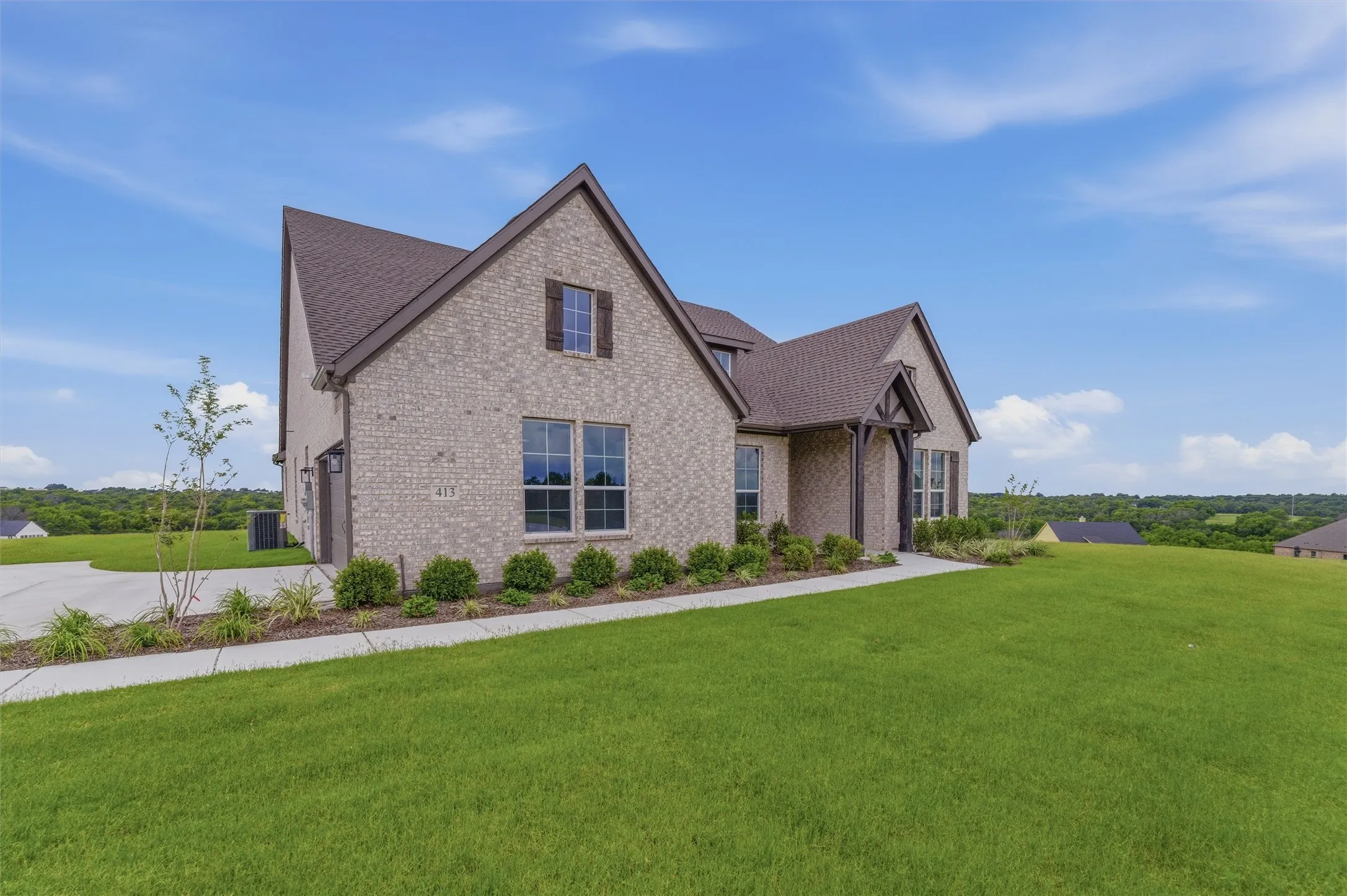 Brick facade residence featuring a prominent gabled roofline, dark brown shutters, a decorative timber frame entry, a concrete driveway, and a well-maintained lawn