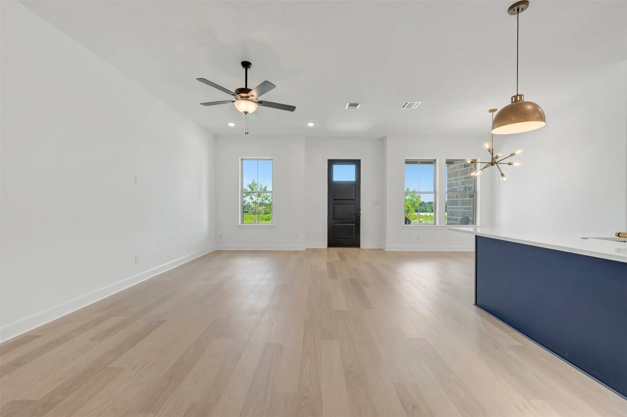 Expansive open-concept living space featuring light wood-finish flooring, crisp white walls, and a contemporary dark blue kitchen island with a white countertop