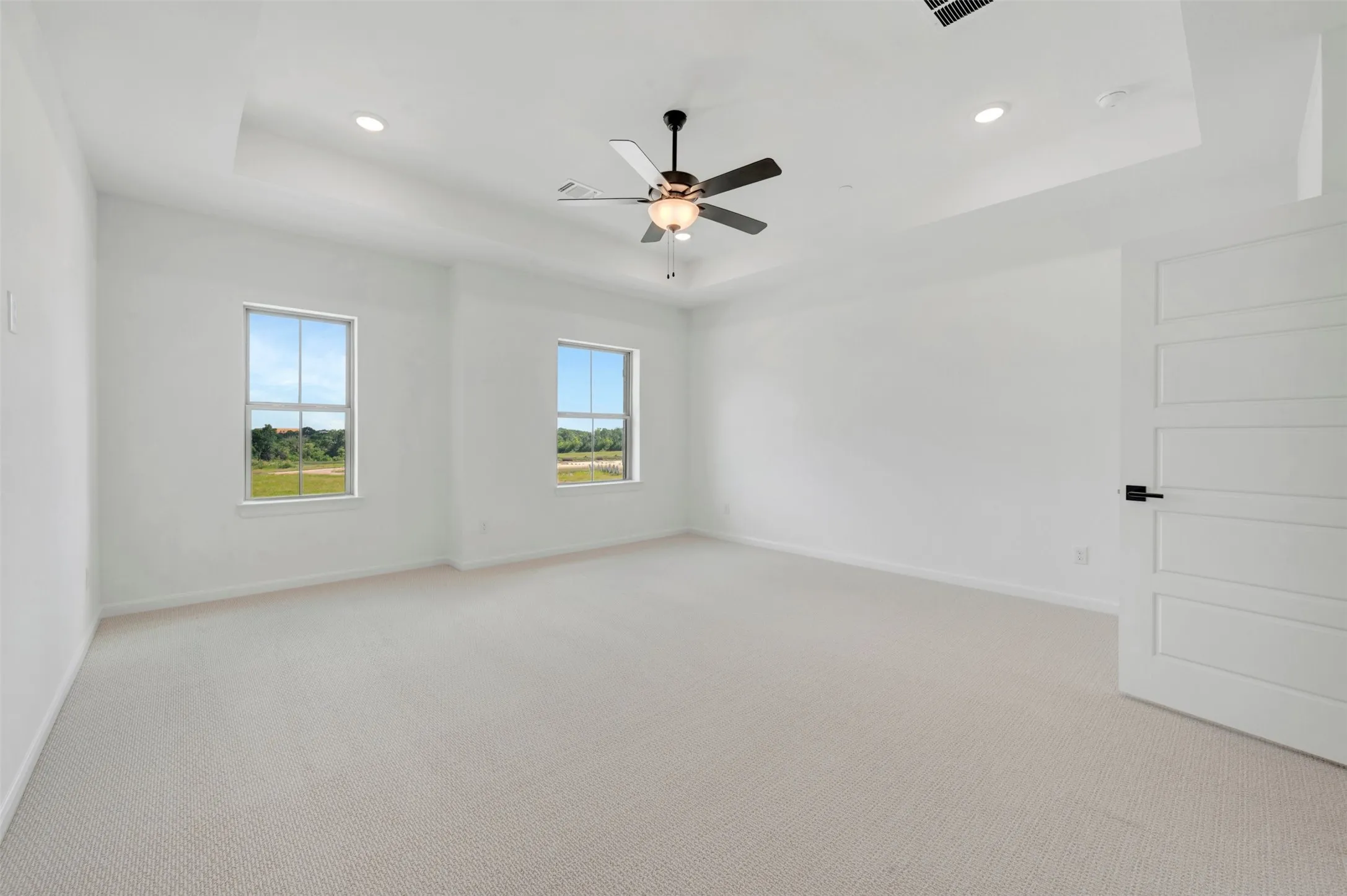 Spacious room featuring a tray ceiling with recessed lighting and a ceiling fan, two double-hung windows, light-toned carpeting, and a modern paneled door with matte black hardware